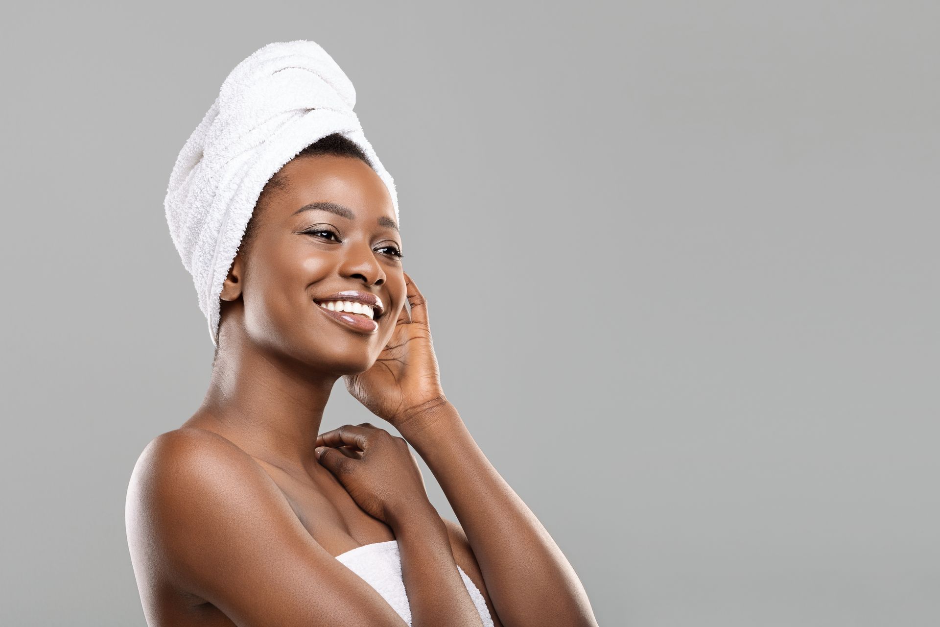 Woman wrapped in a white towel, smiling, with wet skin, against a gray background.