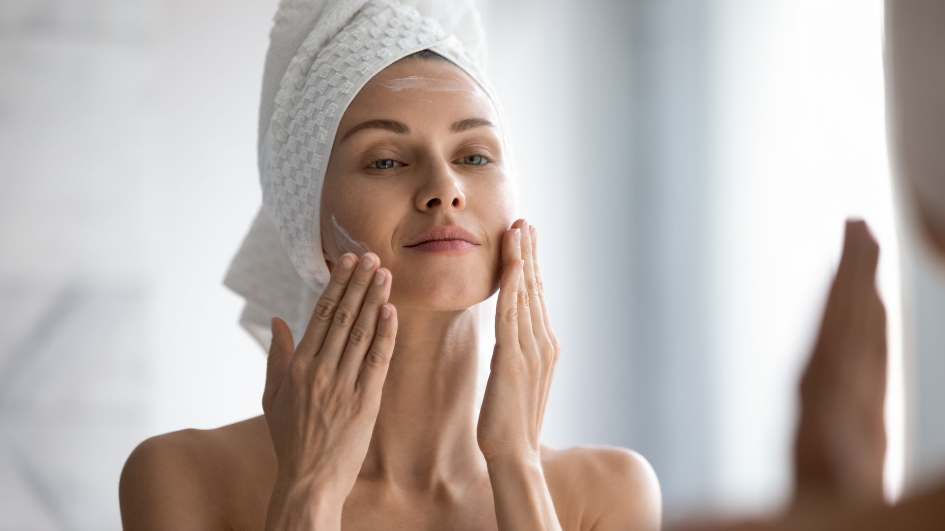 Woman with a towel on her head applies cream to her face in a mirror, looking down, focused, white setting.