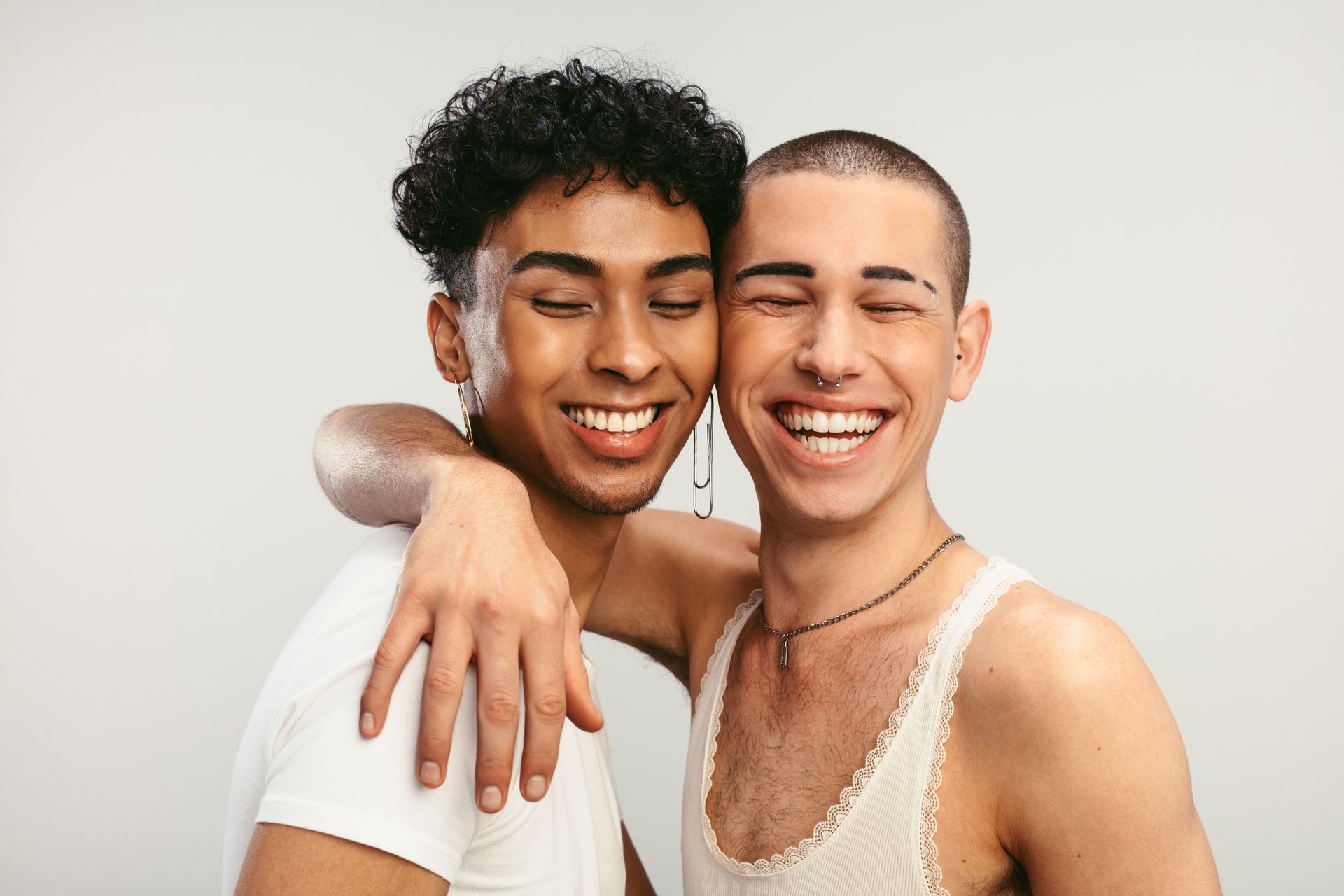 Two smiling people with arms around each other, fair skin, one bald, one with dark curly hair, white background.
