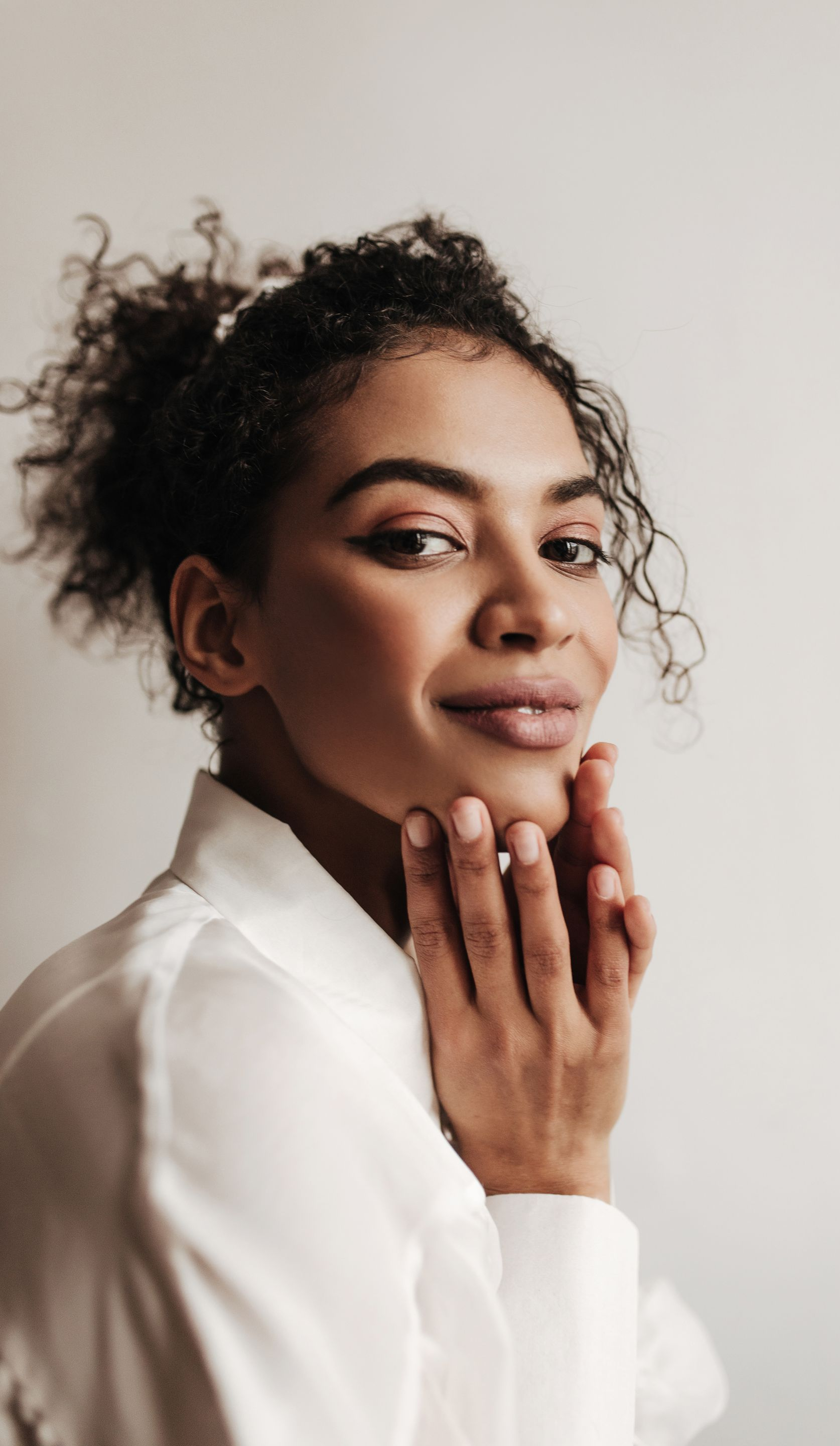 Woman with curly hair in white shirt, smiling and touching her face.