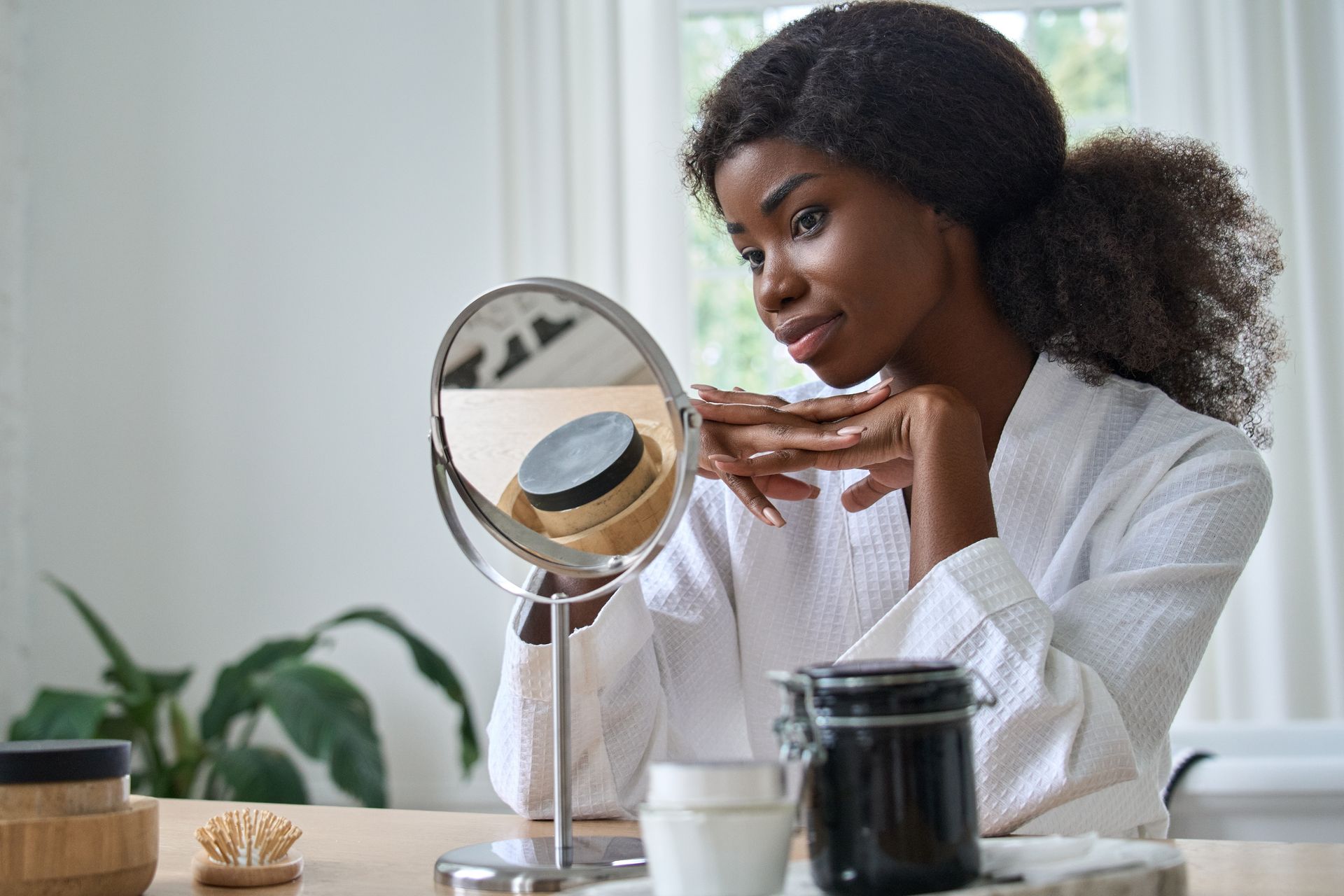 Woman in a white robe looks in a mirror, touching her face in a bright room.