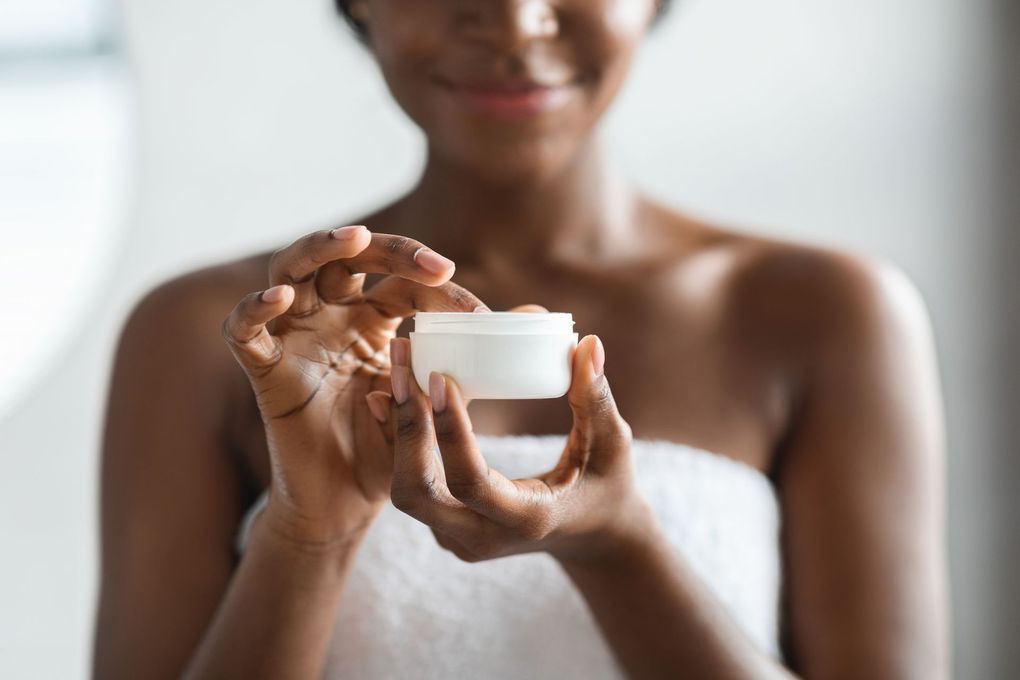 Smiling Black woman holding a white jar of skincare, wrapped in a towel.