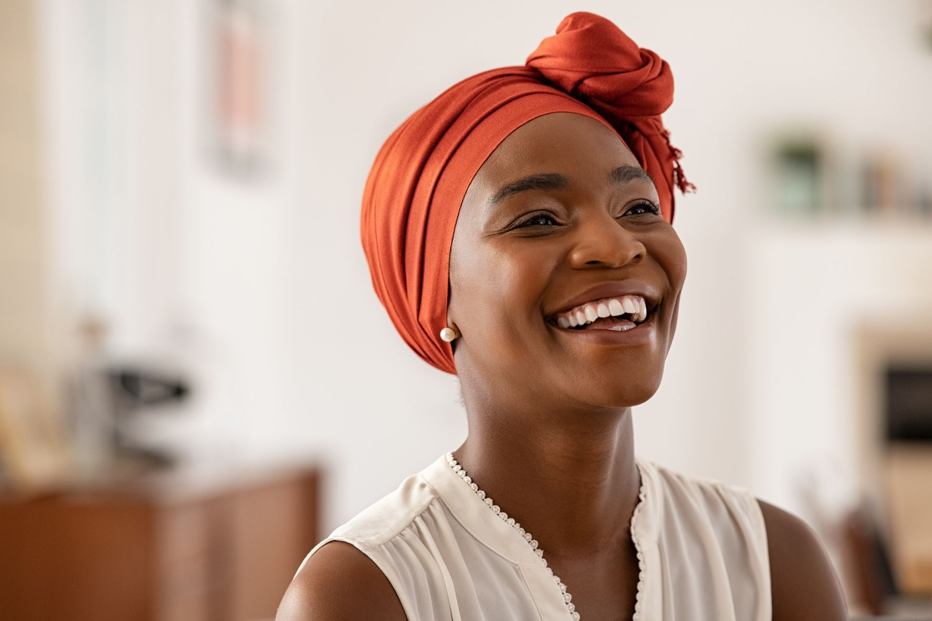 Smiling Black woman wearing an orange headwrap and white top; indoor setting.