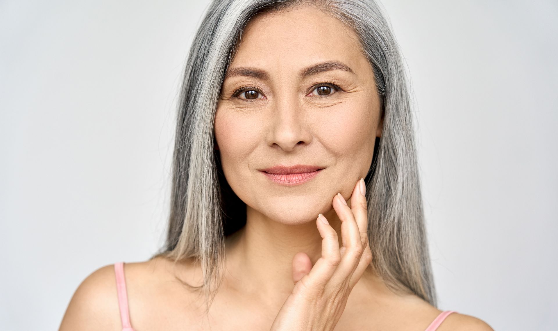 Mature woman with long gray hair, smiling, hand touching her cheek, neutral background.