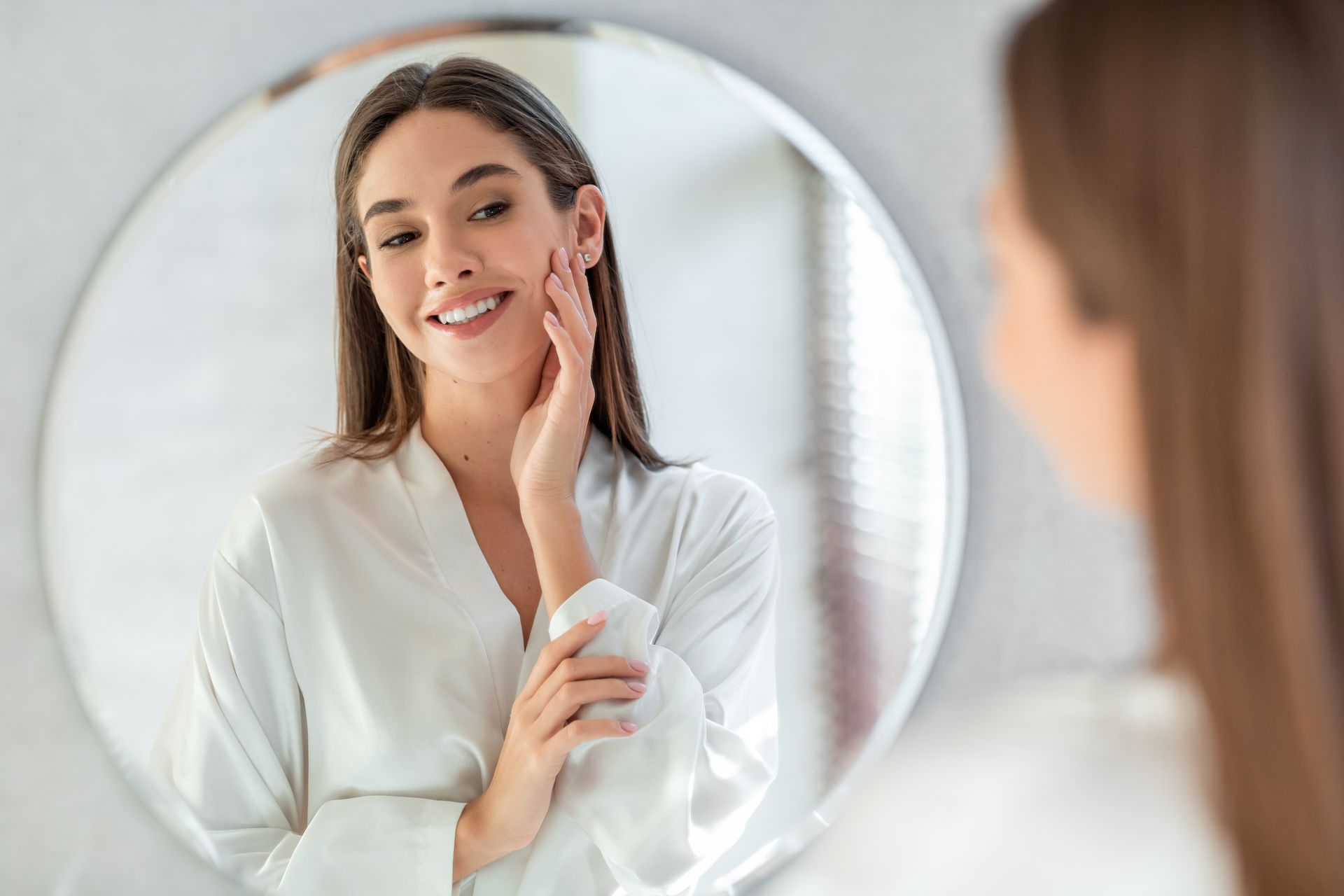Woman in white robe looking in mirror, smiling, touching face.