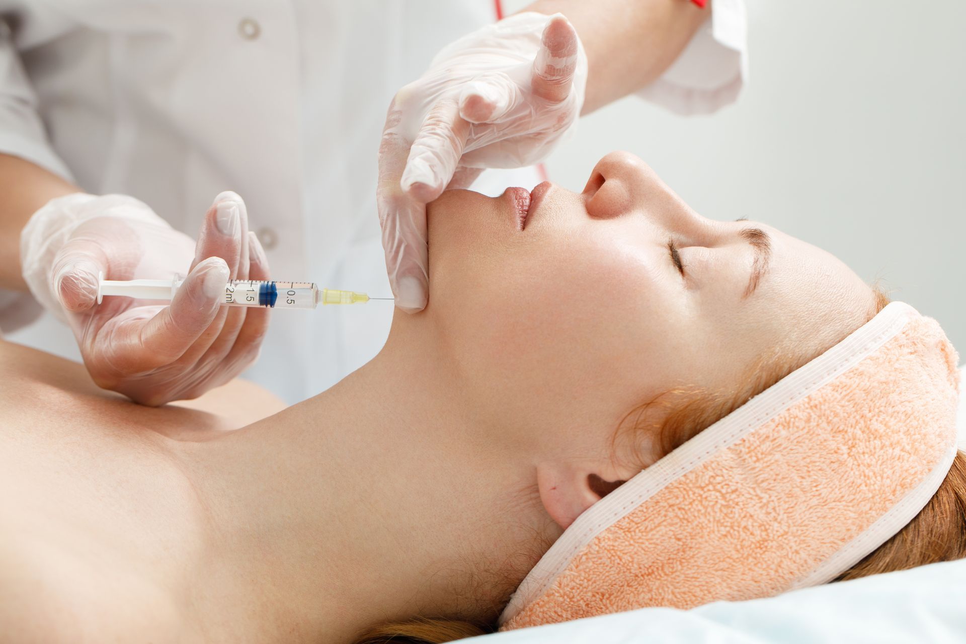 Woman receiving a facial injection, administered by a person wearing gloves. White setting.