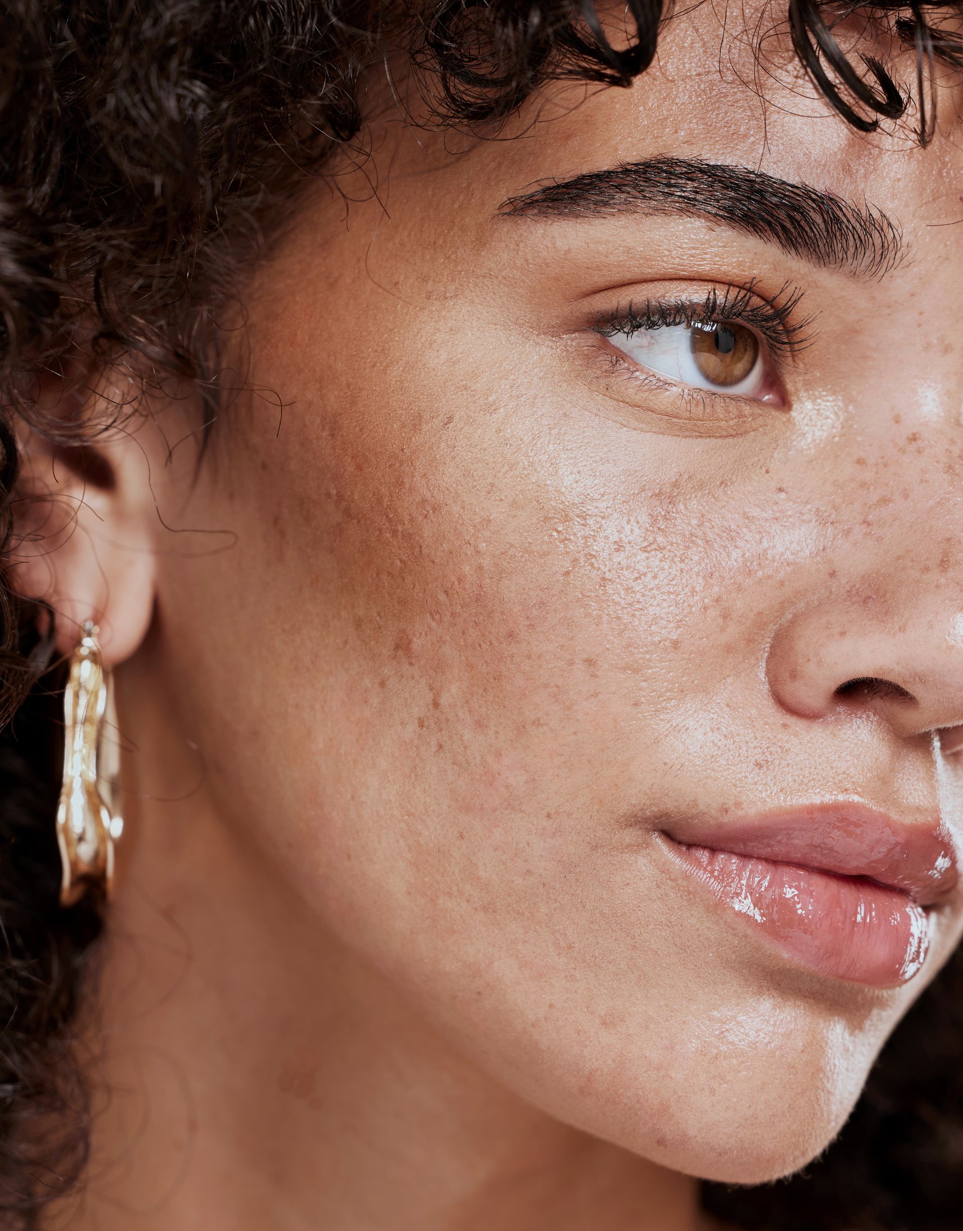 Close-up of a woman with curly hair, freckles, and gold earrings. She has brown eyes and a subtle smile.