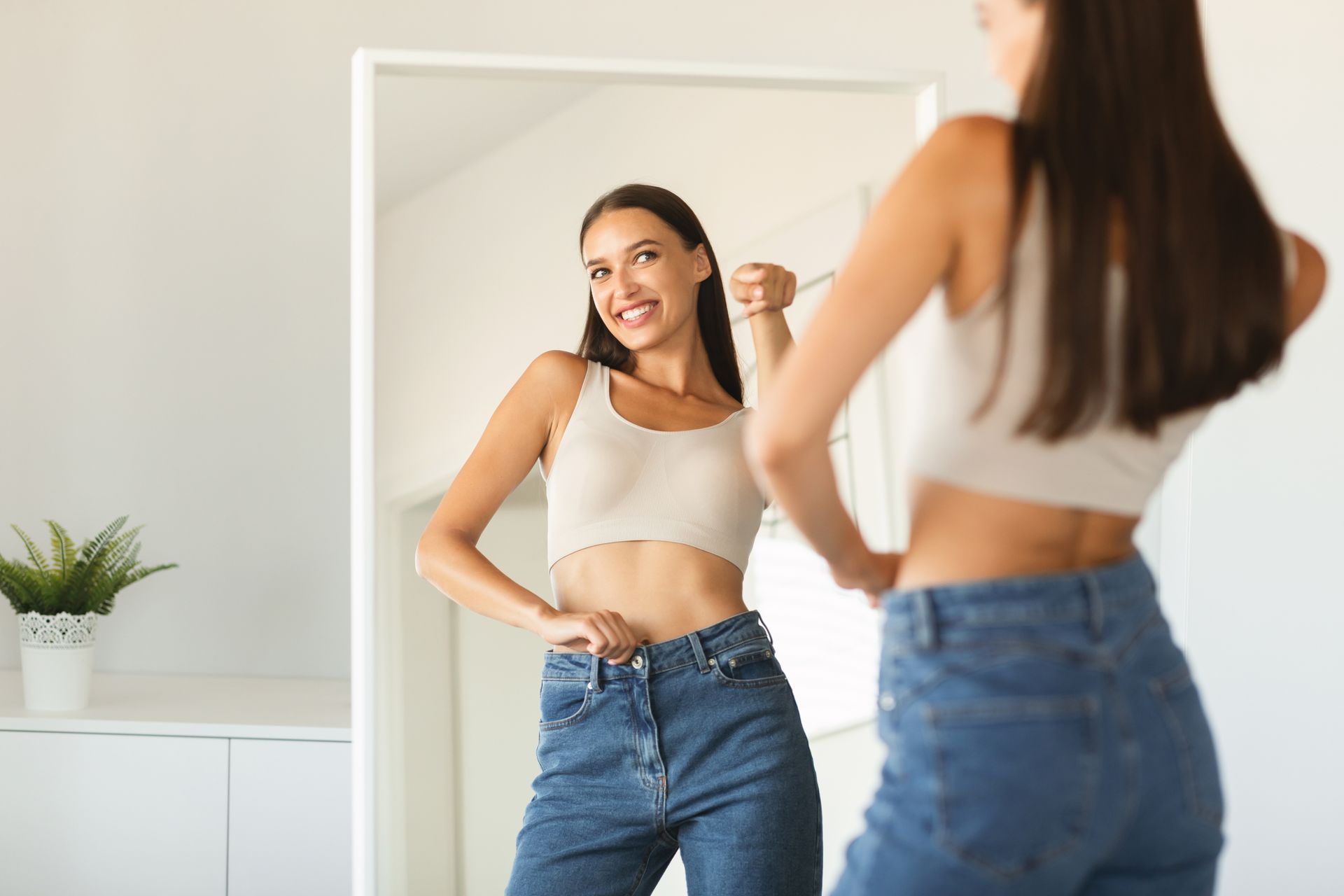 Woman in mirror, smiling, measuring waist with measuring tape, wearing jeans and crop top.