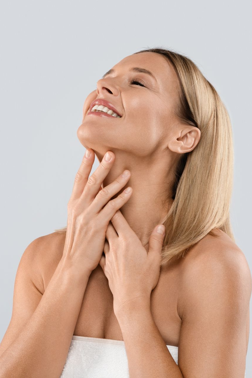 Woman smiling, touching her neck with both hands, eyes closed, wrapped in white towel.