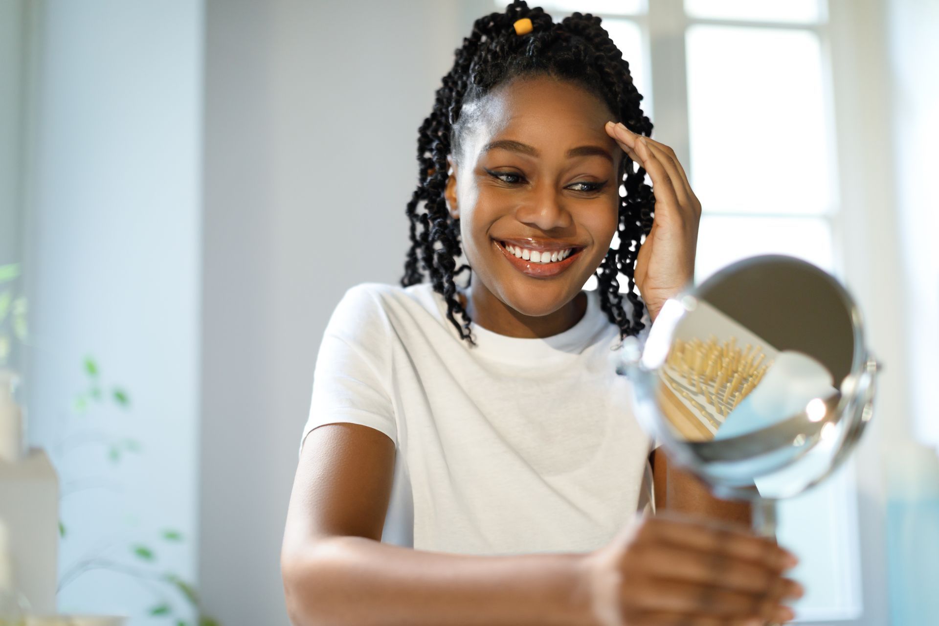 Smiling Black woman examining face in a small mirror, indoors, white t-shirt, window light.