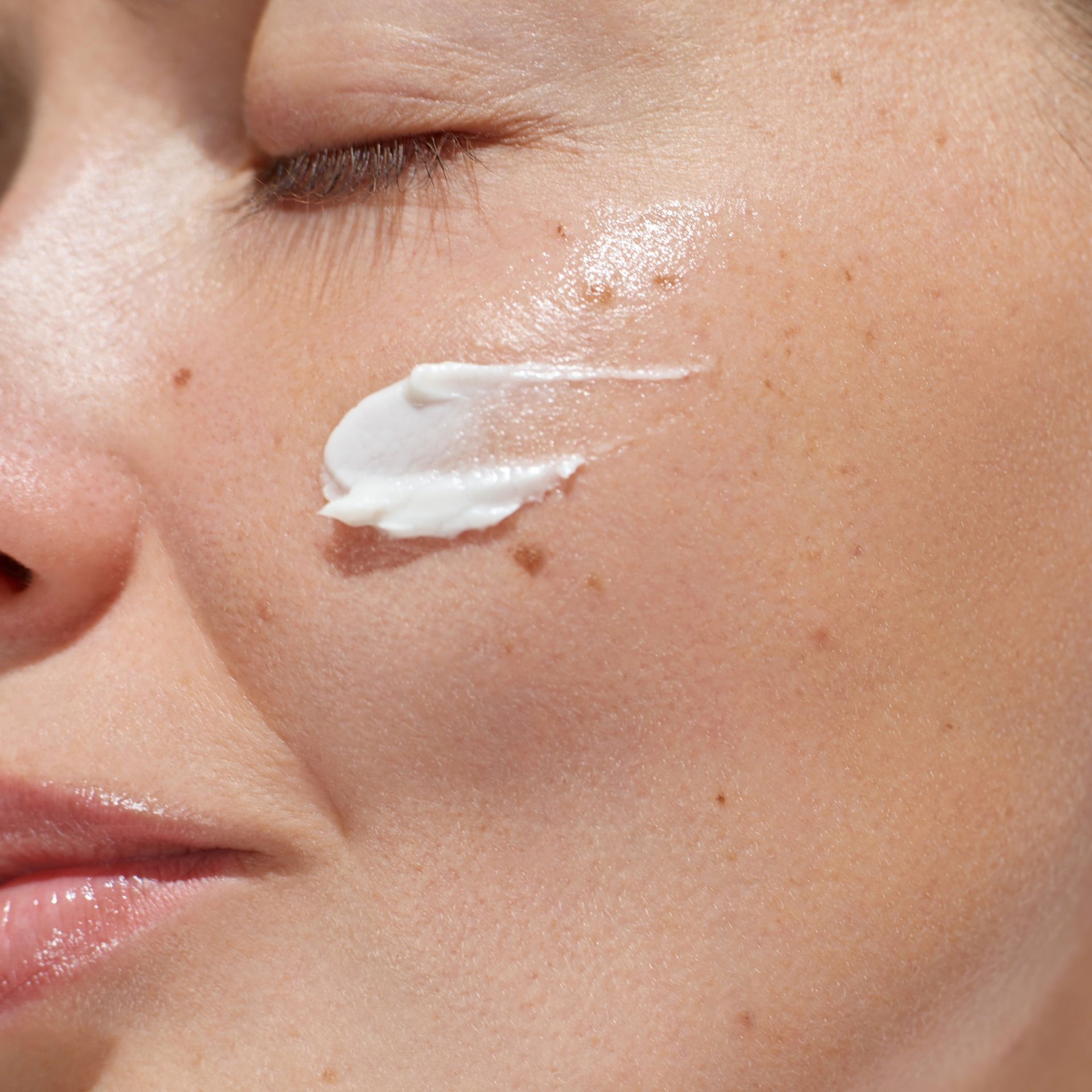 A close-up of a person applying white cream to their cheek with closed eyes.