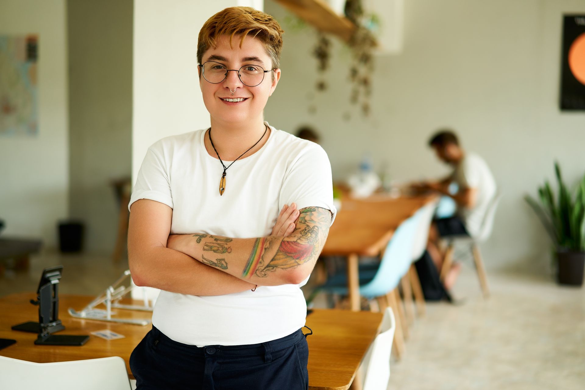 Person with short hair, glasses, and arm tattoos smiles with arms crossed in an office setting.