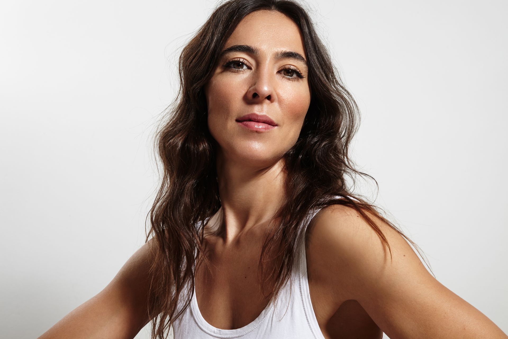 Woman with dark wavy hair, wearing a white tank top, smiling at camera, studio background.