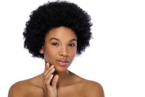 Woman with dark skin and afro touching her cheek, looking at the camera with a neutral expression, on a white background.