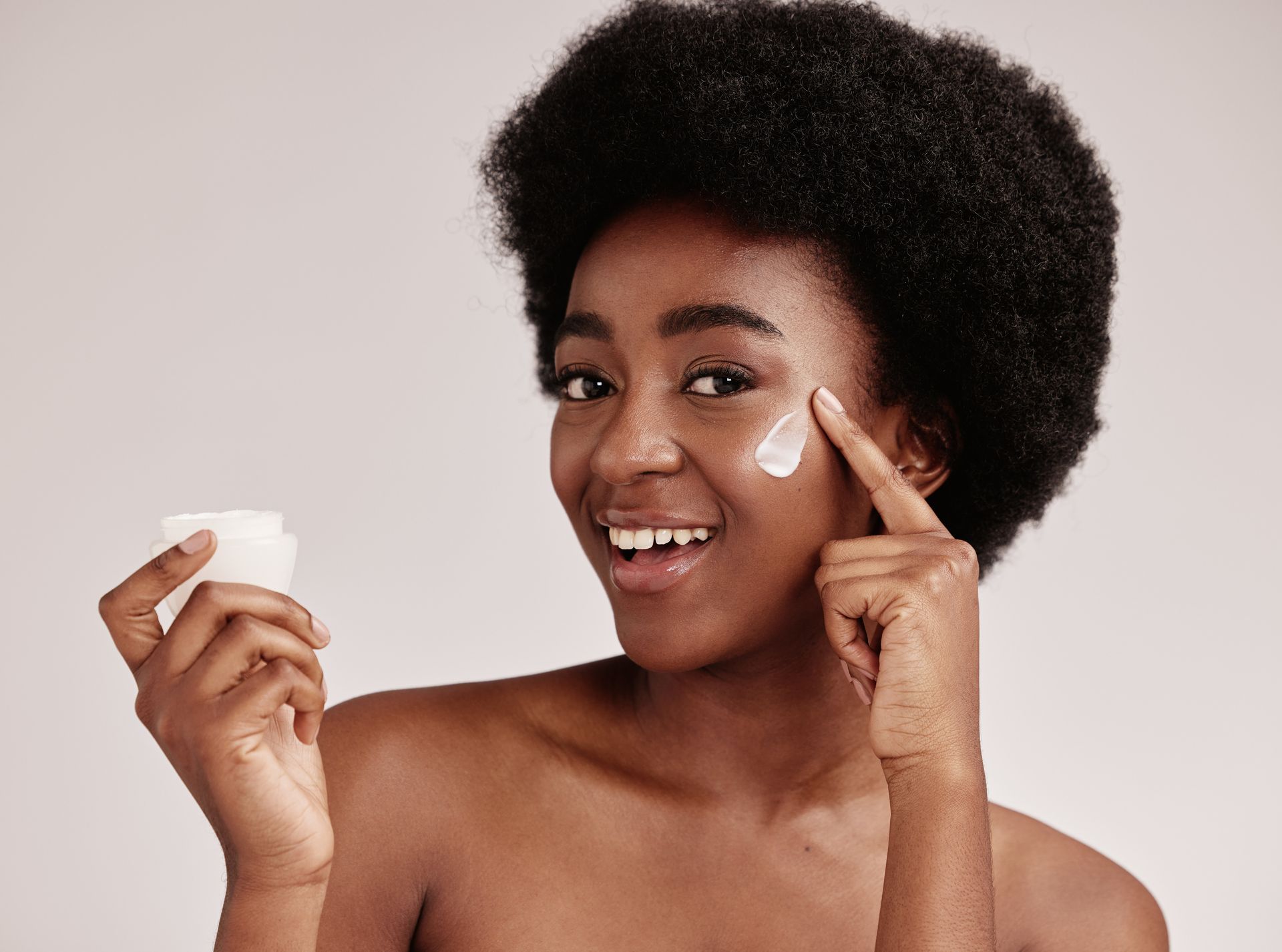 Smiling Black woman applying skincare cream to her face, holding a small jar, against a neutral backdrop.