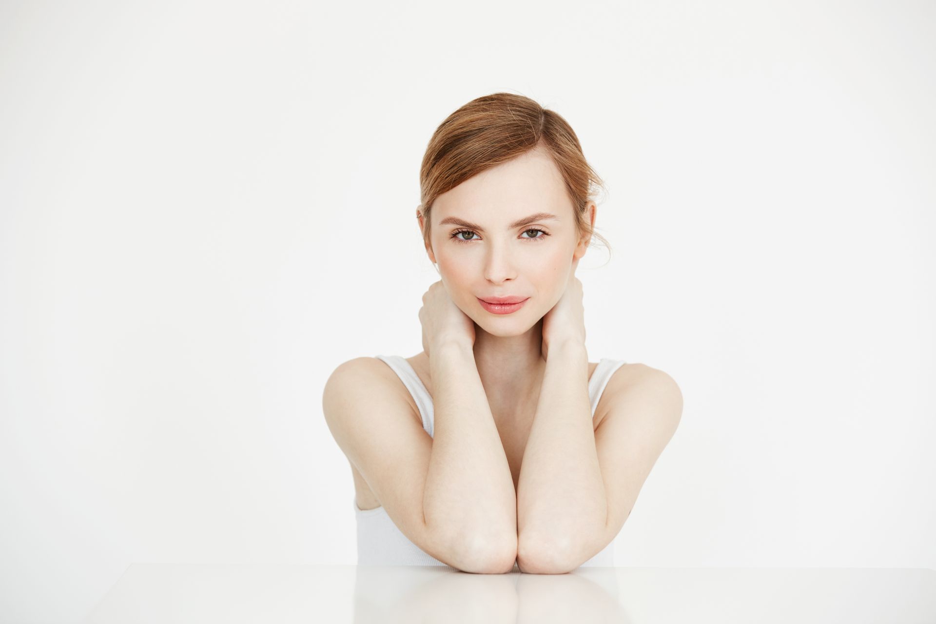 Woman with reddish hair in a white tank top, hands near her neck, looking at camera. White background.
