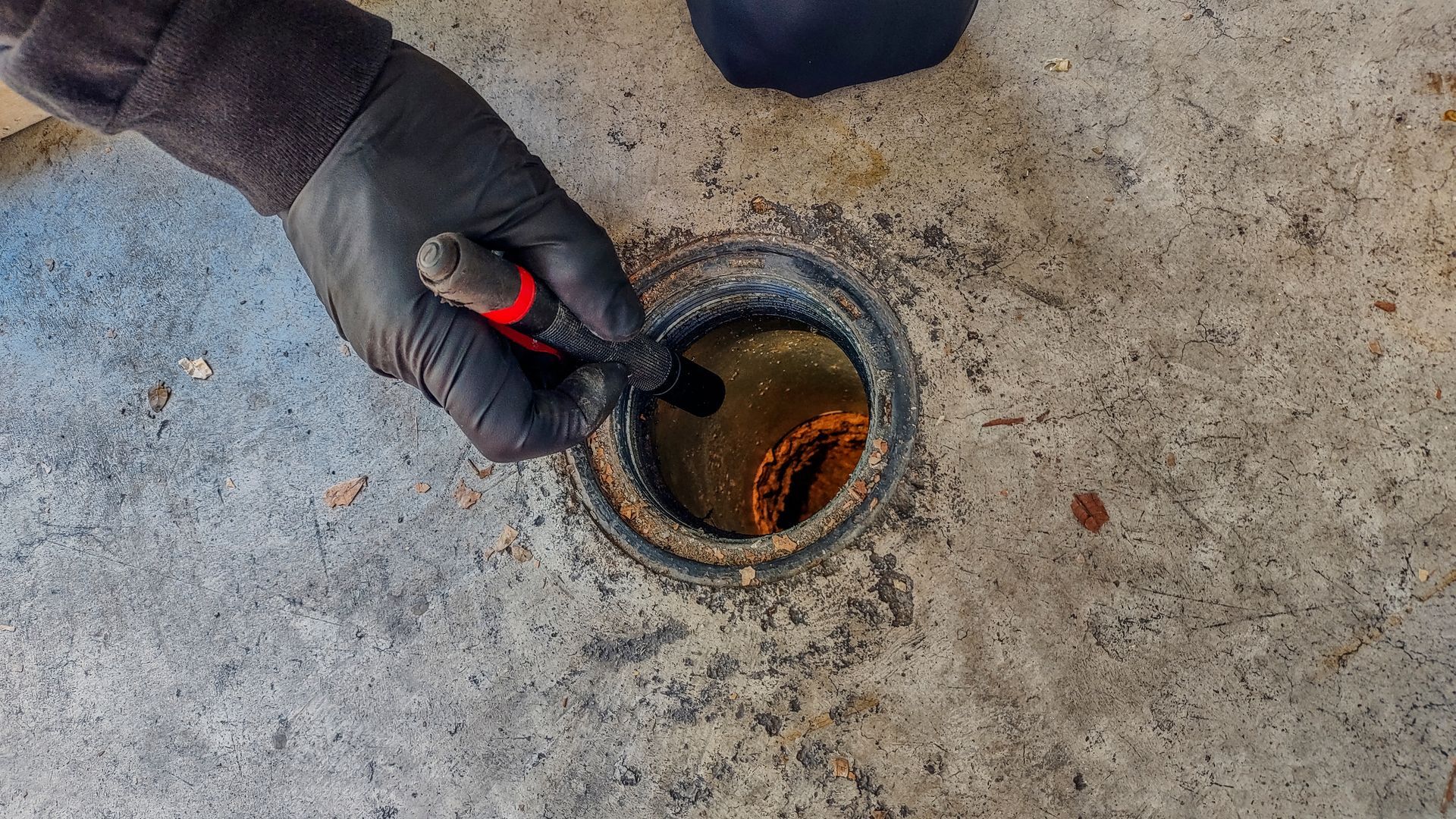 Gloved hand using a tool to inspect a round floor drain in a concrete surface