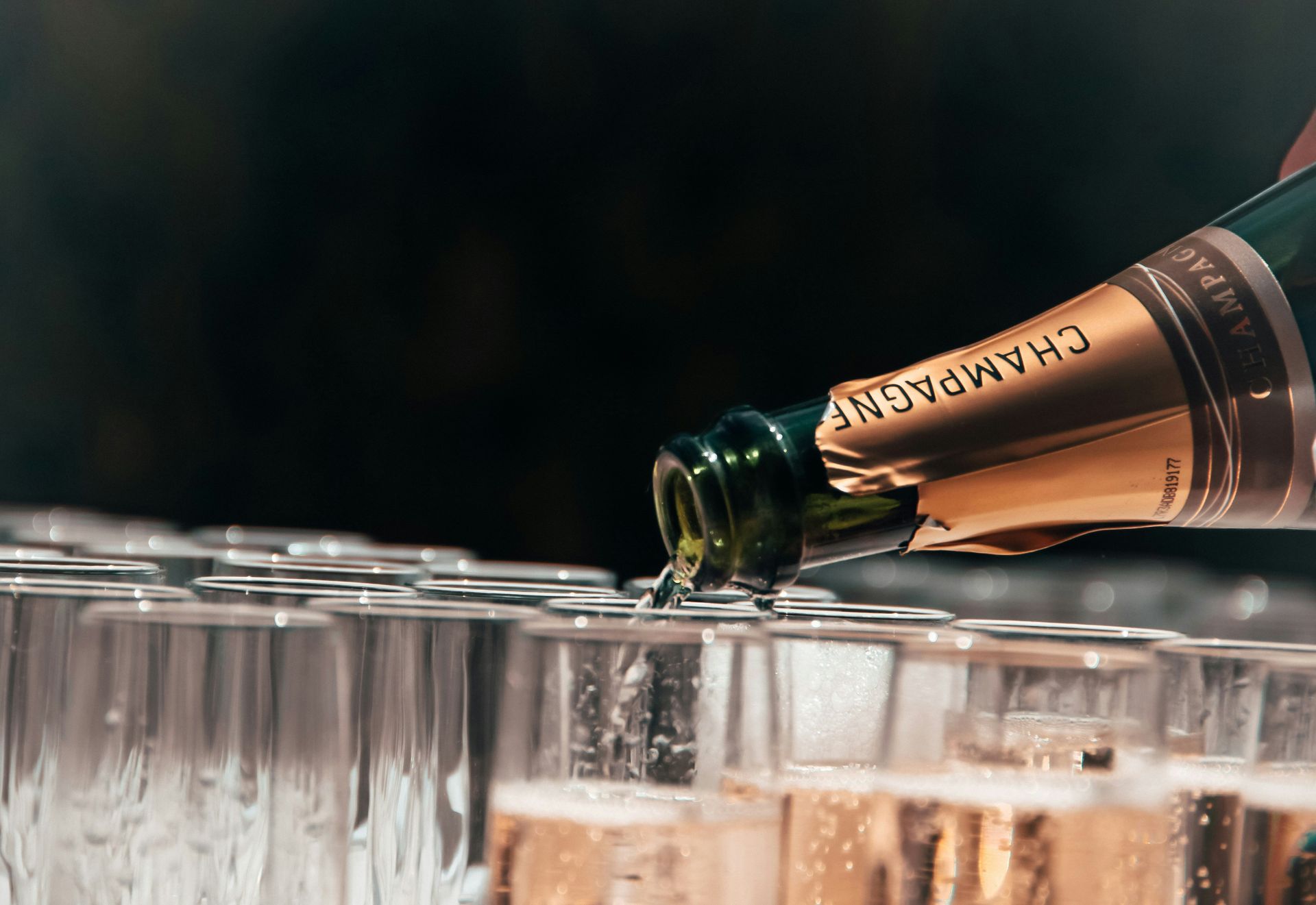 A bottle of champagne is being poured into a row of champagne flutes.