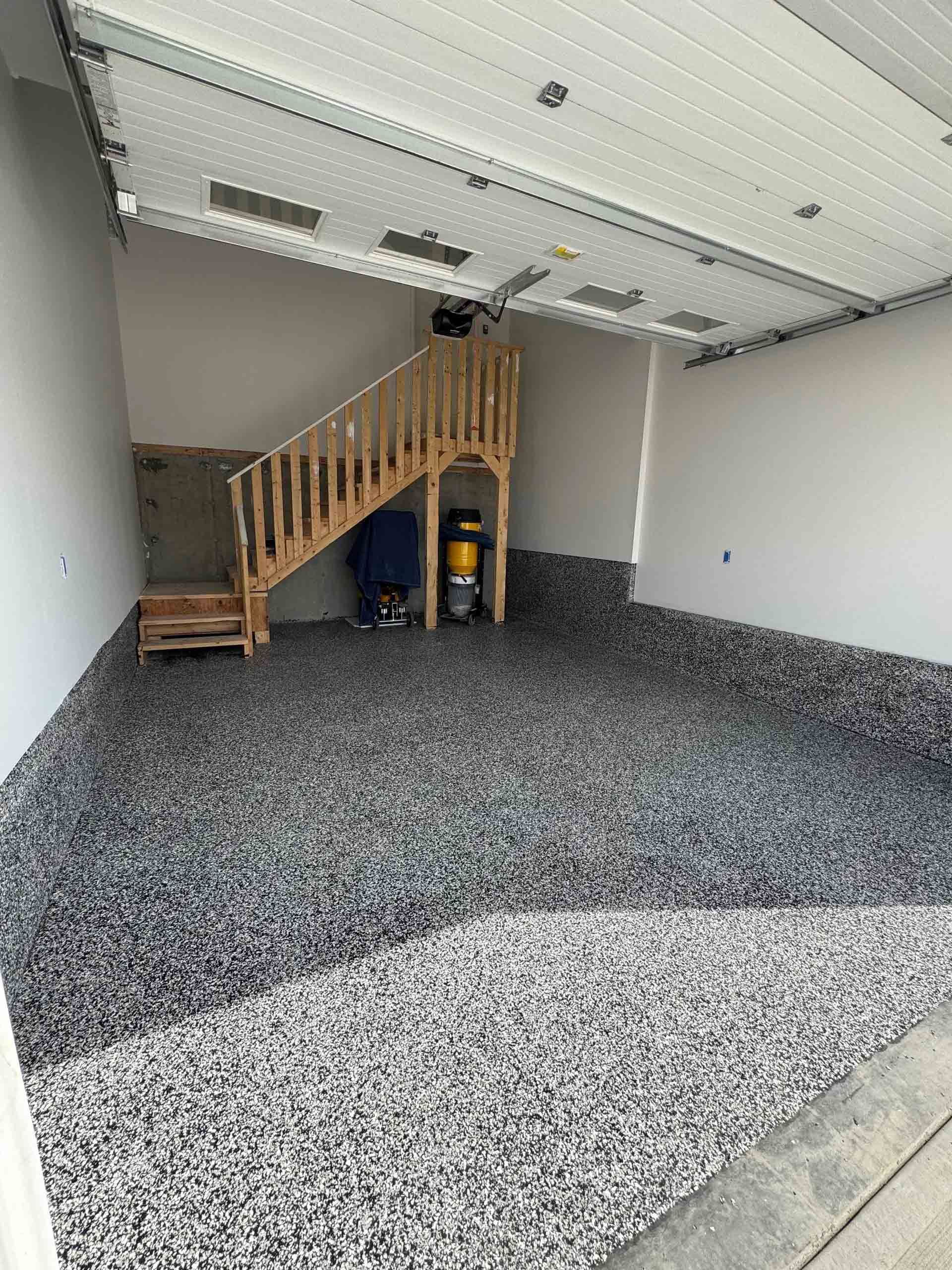 Empty garage with speckled epoxy floor and a wooden staircase leading to a loft area.