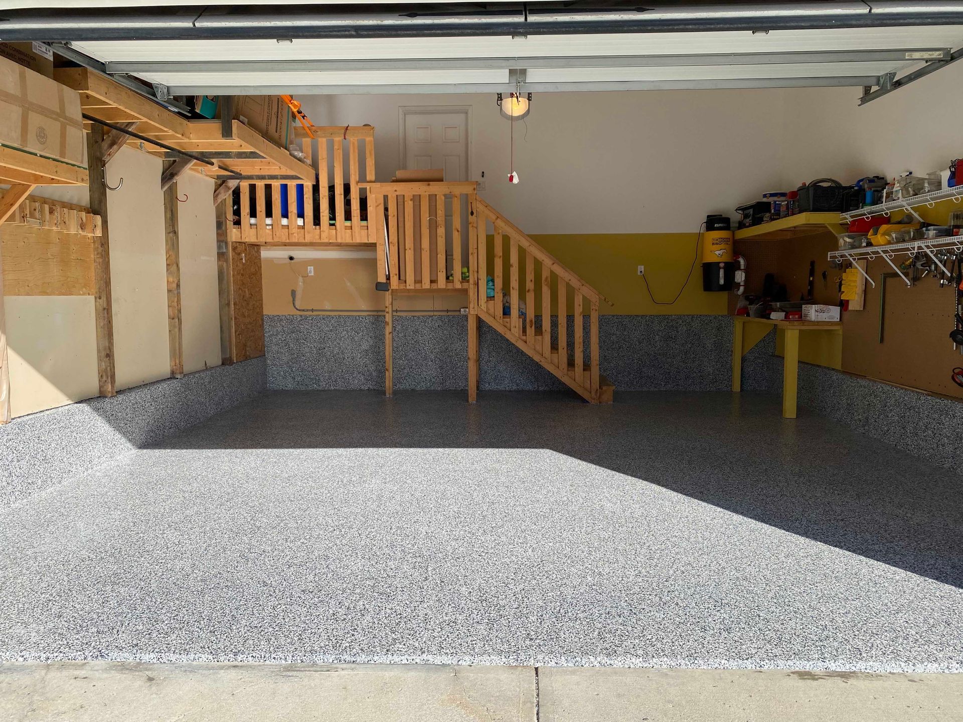 Empty garage with a speckled epoxy floor, wooden stairs to a loft, and shelves along the right wall