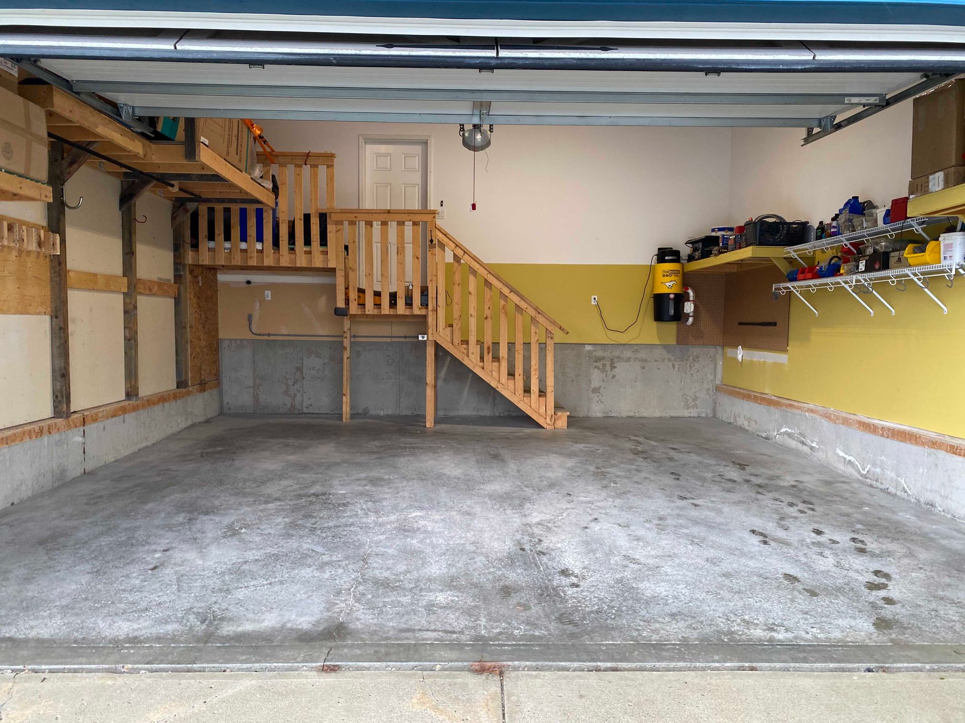 Empty garage with concrete floor, wood staircase to a loft, and yellow walls with shelves along the right side.