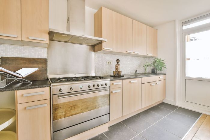 Bright modern kitchen with light wood cabinets, stainless steel stove, gray floor, and a sunny door.