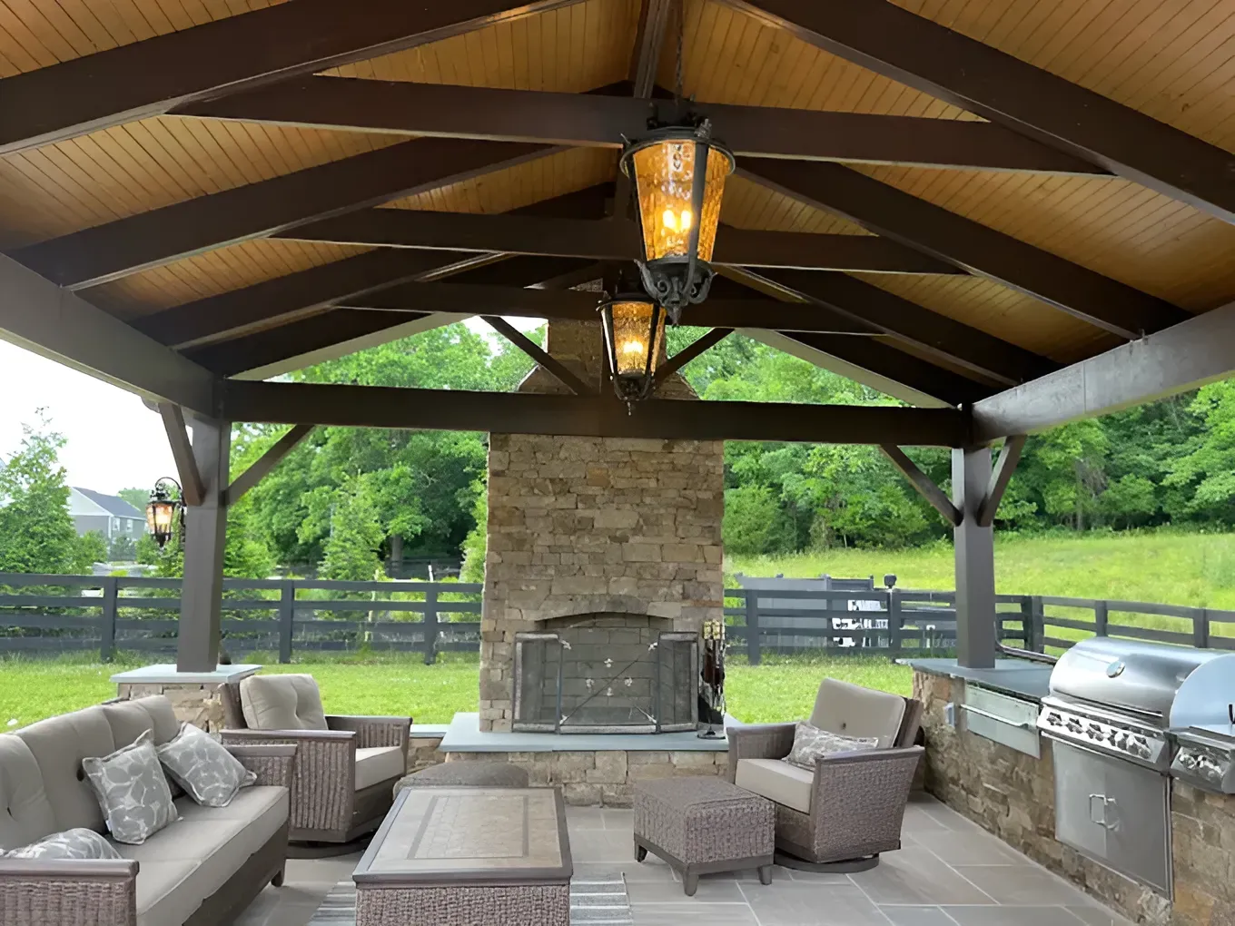 Covered outdoor patio with wooden beams, stone fireplace, wicker seating, and a built-in grill beneath a peaked roof.