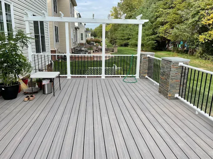 Back deck with gray composite flooring, white pergola, railing, and potted plants beside a house
