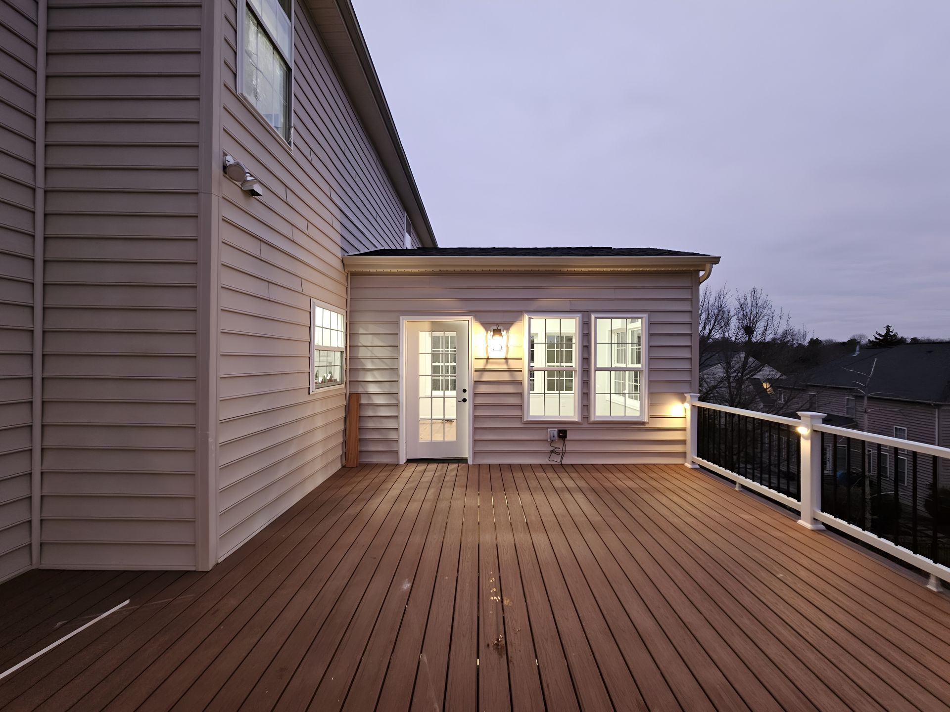 Deck at dusk beside a house, with lit windows and a railing overlooking water.