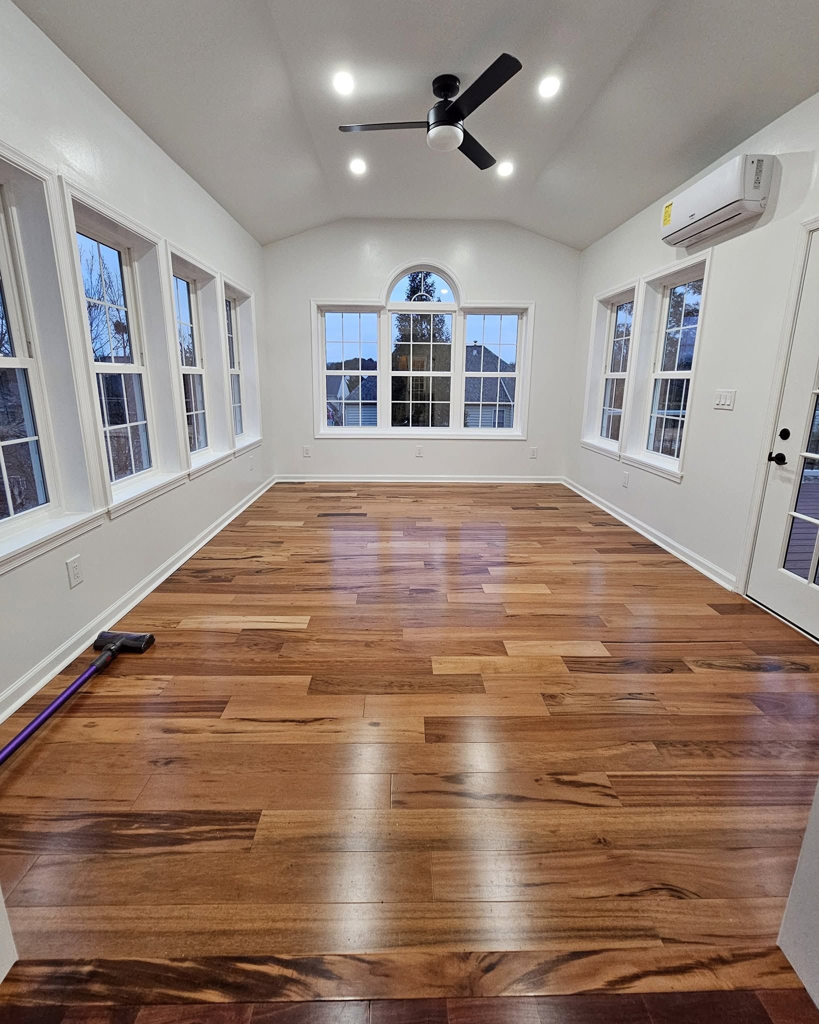 Bright empty room with hardwood floors, white walls, ceiling fan, and large windows.