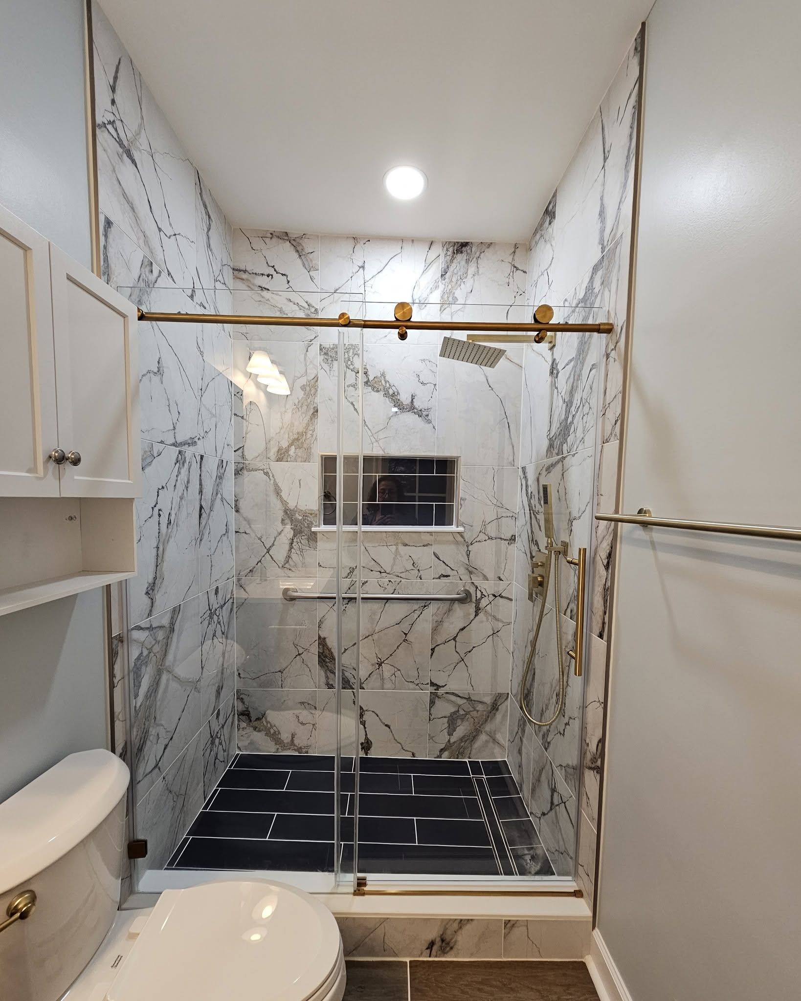 Bathroom with marble shower stall, black tile floor, gold grab bars, and toilet on the left
