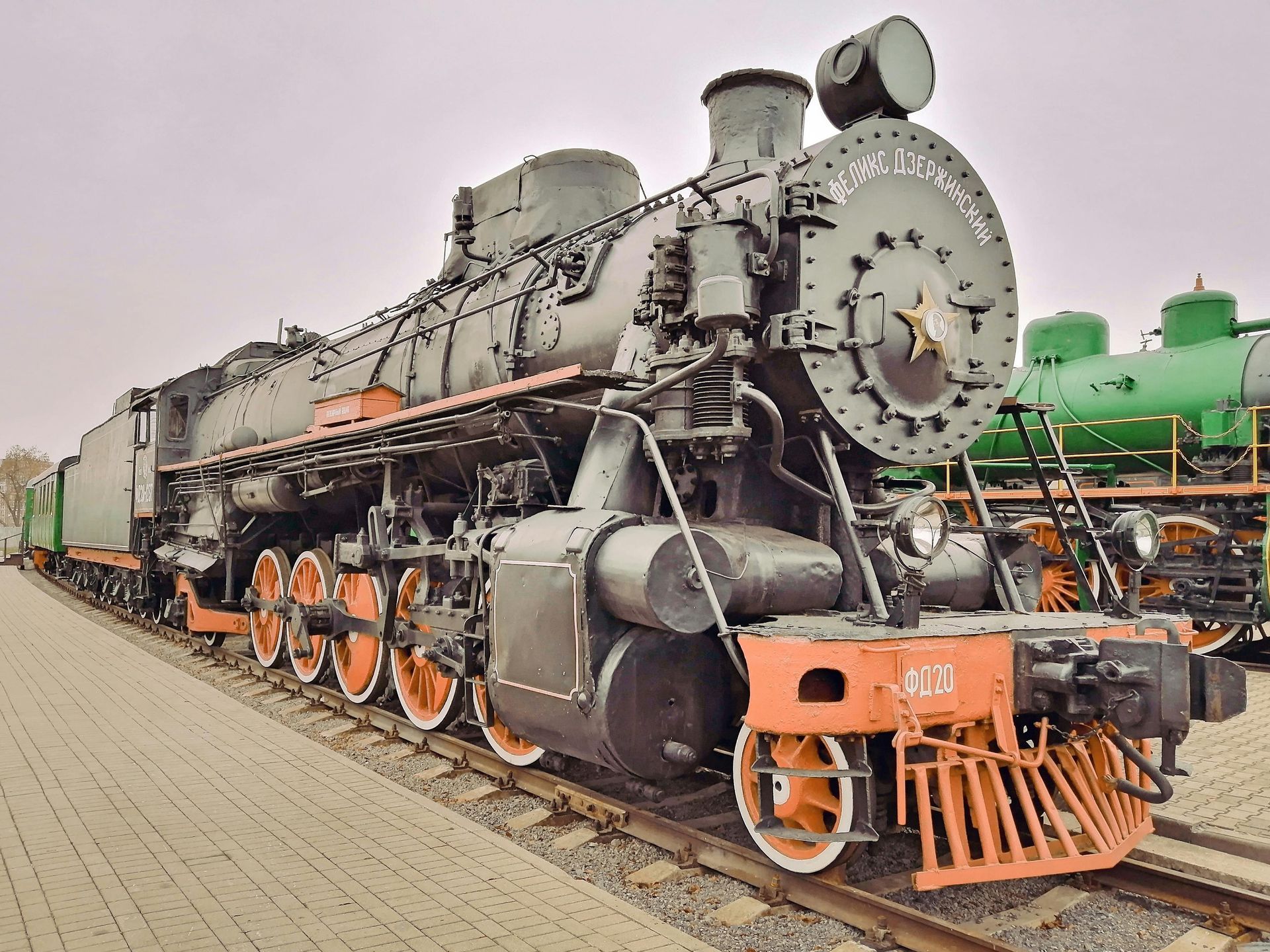 Een zwarte en oranje stoomlocomotief op de rails, met daarachter een groene wagon, in de buitenlucht.