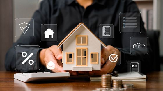 Person holding a model house with financial and real estate icons; calculator and coins on table. Person holding a model house with financial and real estate icons; calculator and coins on table.