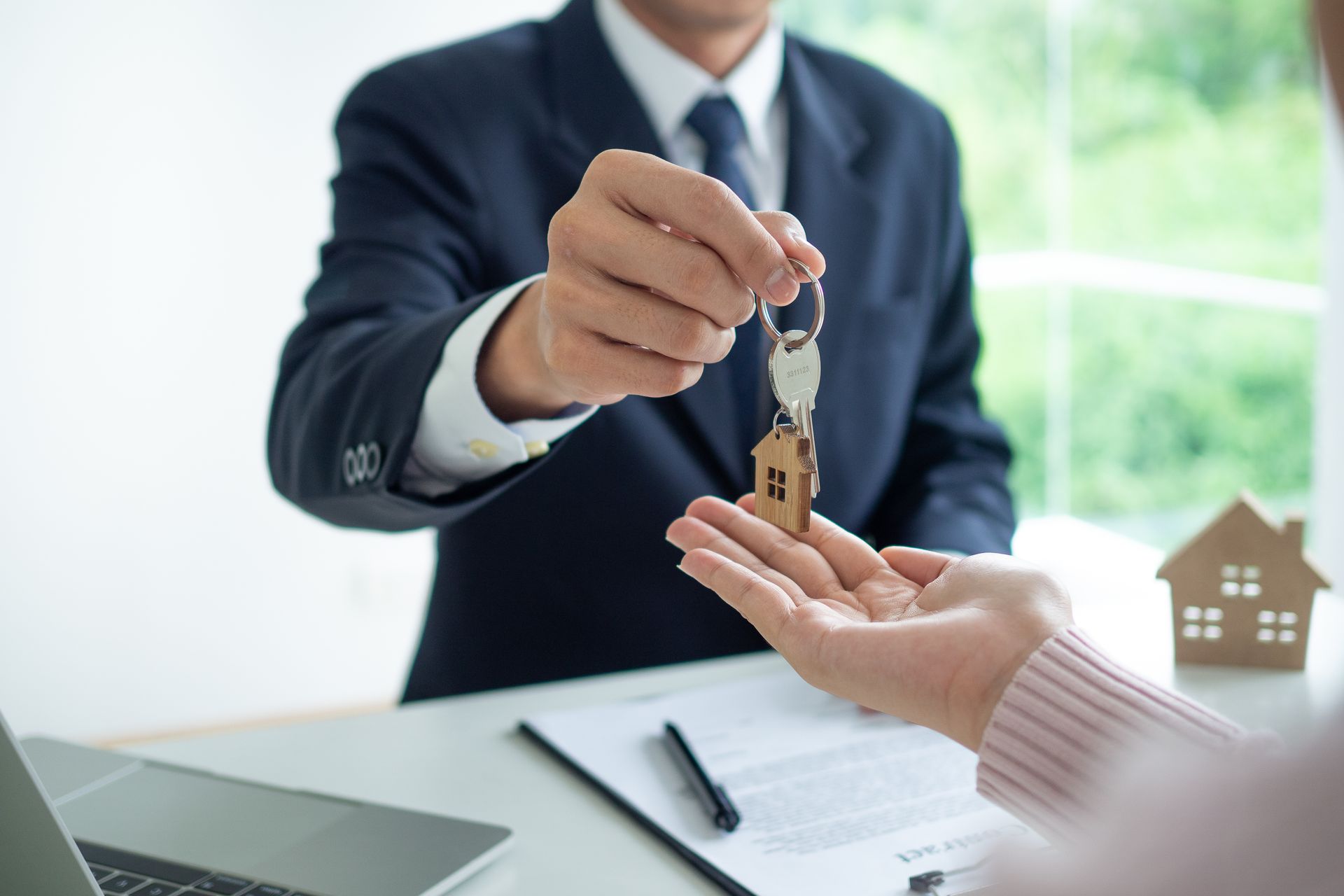 Person in suit handing keys to another person over a desk. A h ouse model and paperwork are visible. Person in suit handing keys to another person over a desk. A h ouse model and paperwork are visible.