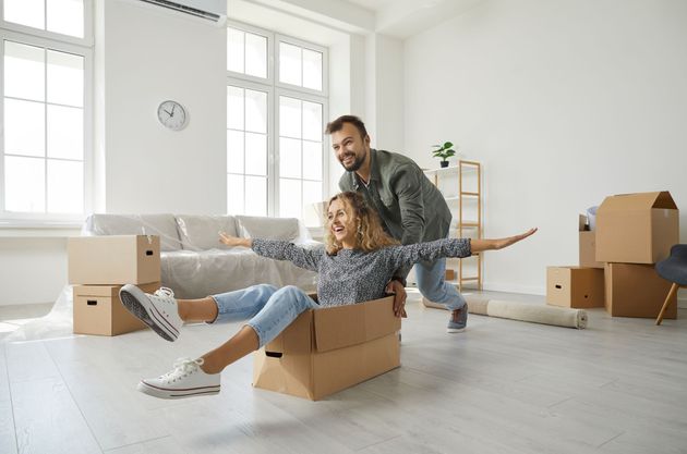 Man pushing woman in a cardboard box, arms outstretched, in a new home with boxes and a sofa. Man pushing woman in a cardboard box, arms outstretched, in a new home with boxes and a sofa.