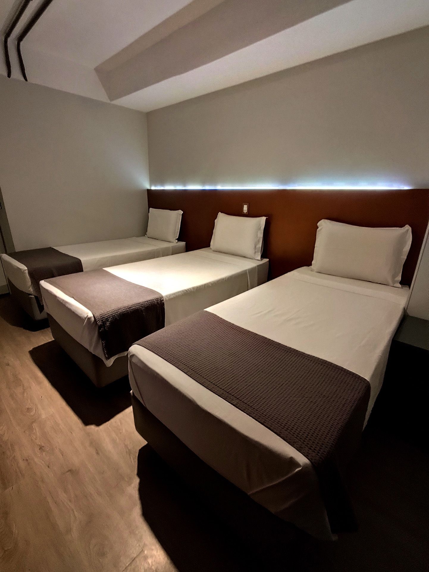Três camas de solteiro em um quarto de hotel com lençóis brancos, colchas marrons e cabeceiras de madeira.