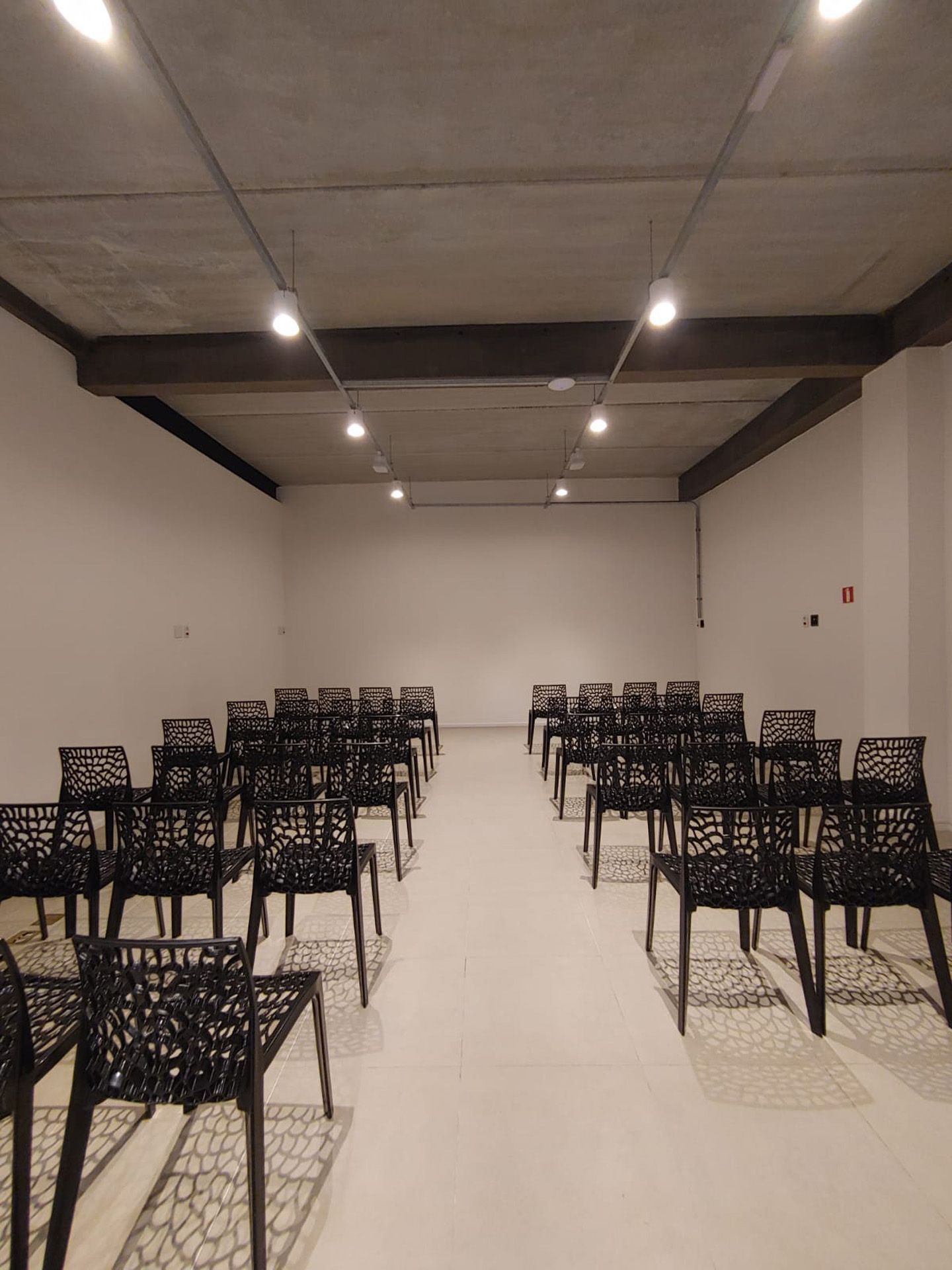 Sala de reuniões vazia com cadeiras pretas dispostas em fileiras, paredes brancas e teto de concreto.