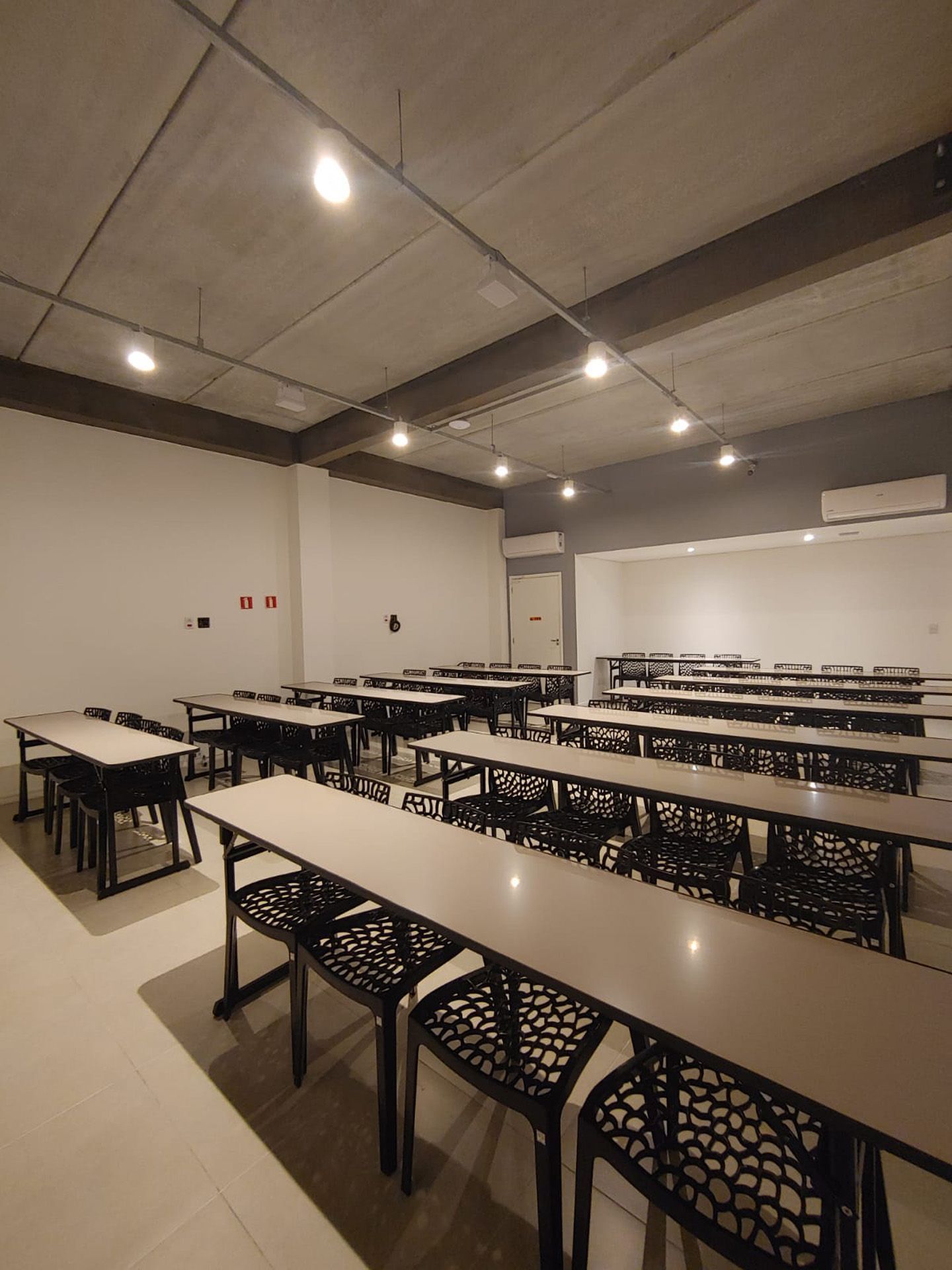Sala de aula vazia com fileiras de mesas e cadeiras; teto de concreto, cores neutras.