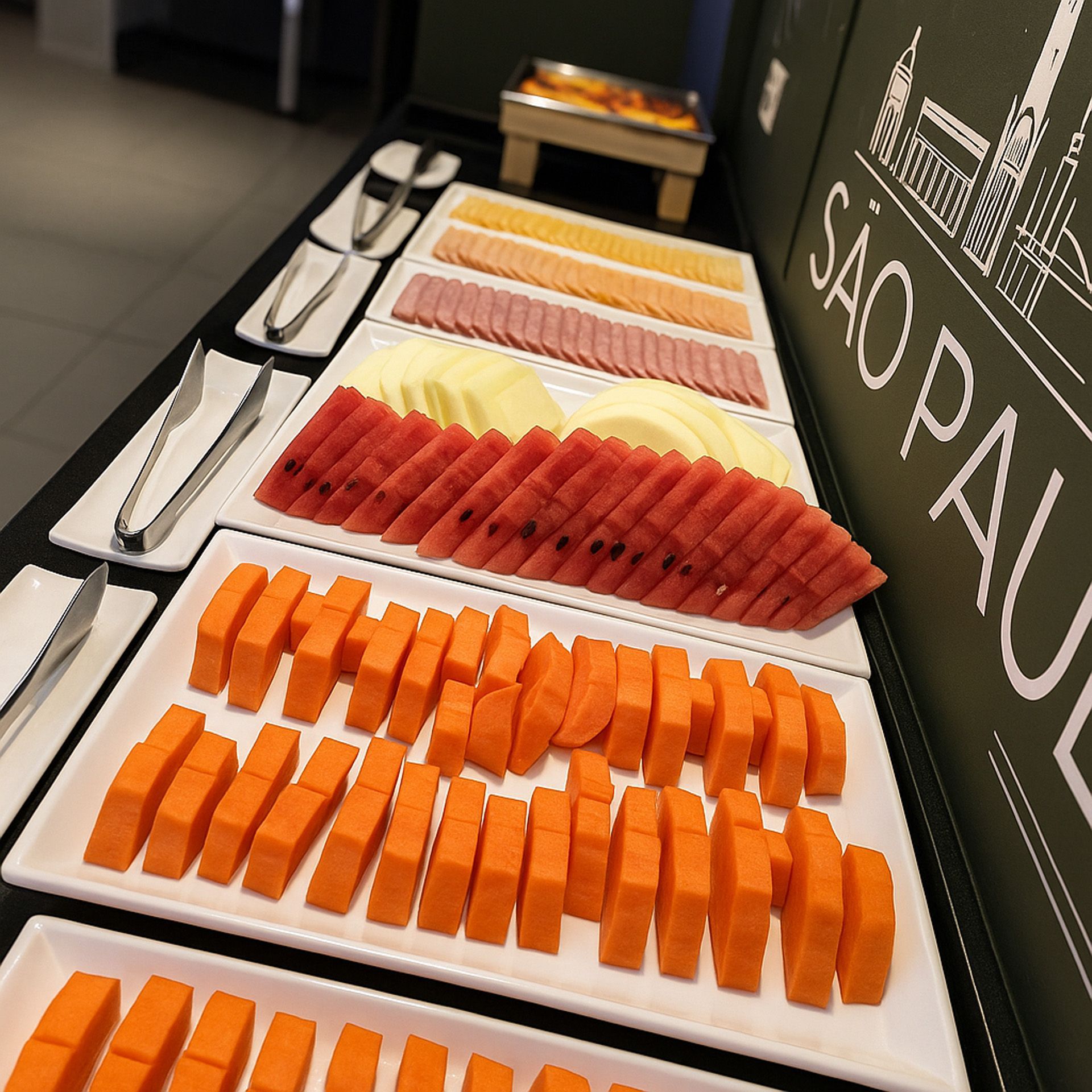 Buffet com frutas fatiadas e frios, servidos em travessas brancas. Placa de São Paulo à direita.