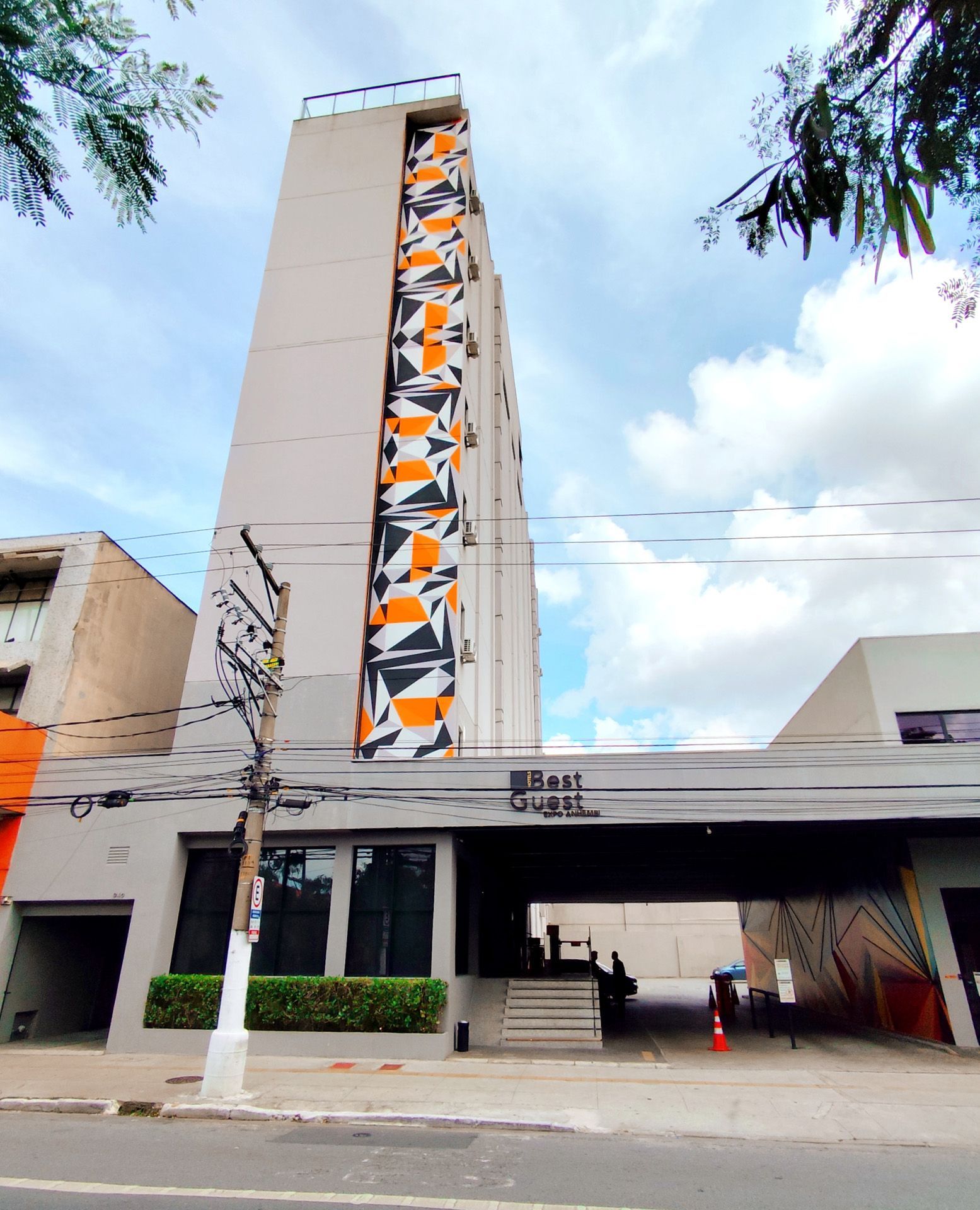 Prédio alto com padrão geométrico laranja e preto, fachada cinza, sob um céu nublado.