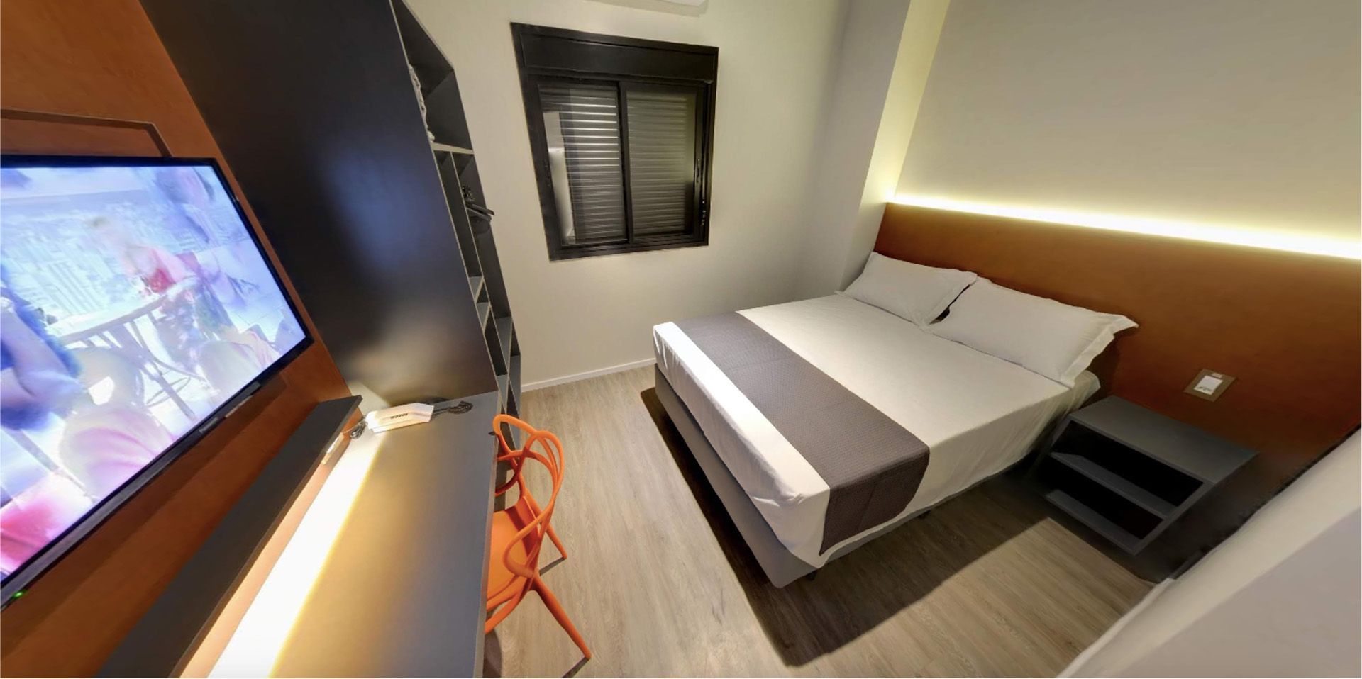 Quarto com cama, TV, cadeira laranja e janela.
