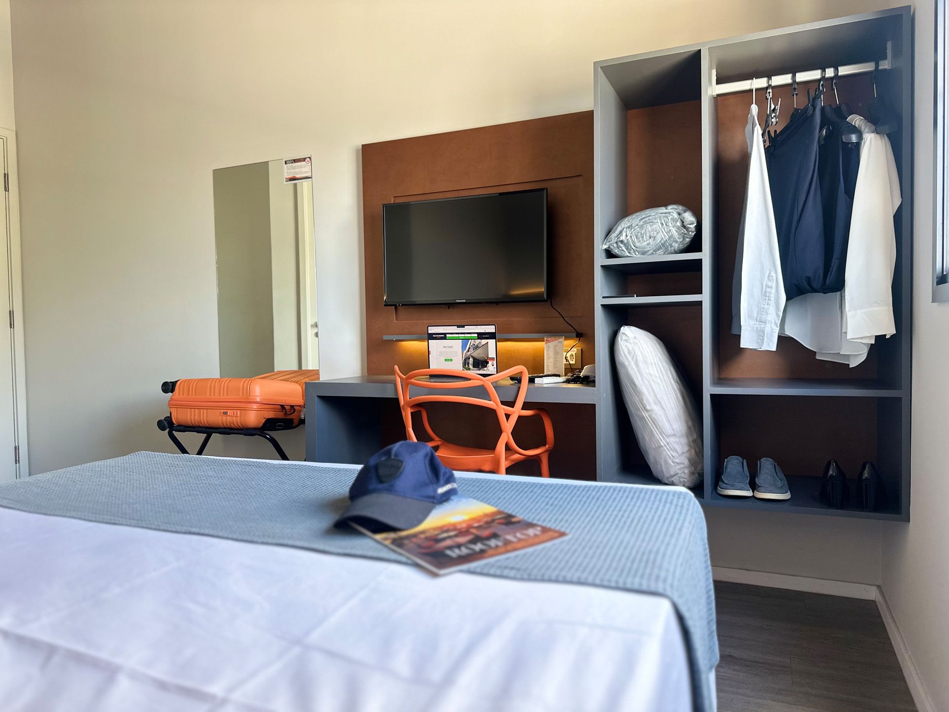 Quarto de hotel com cama, escrivaninha, TV, armário e espaço para bagagem. Tons de laranja e cinza.