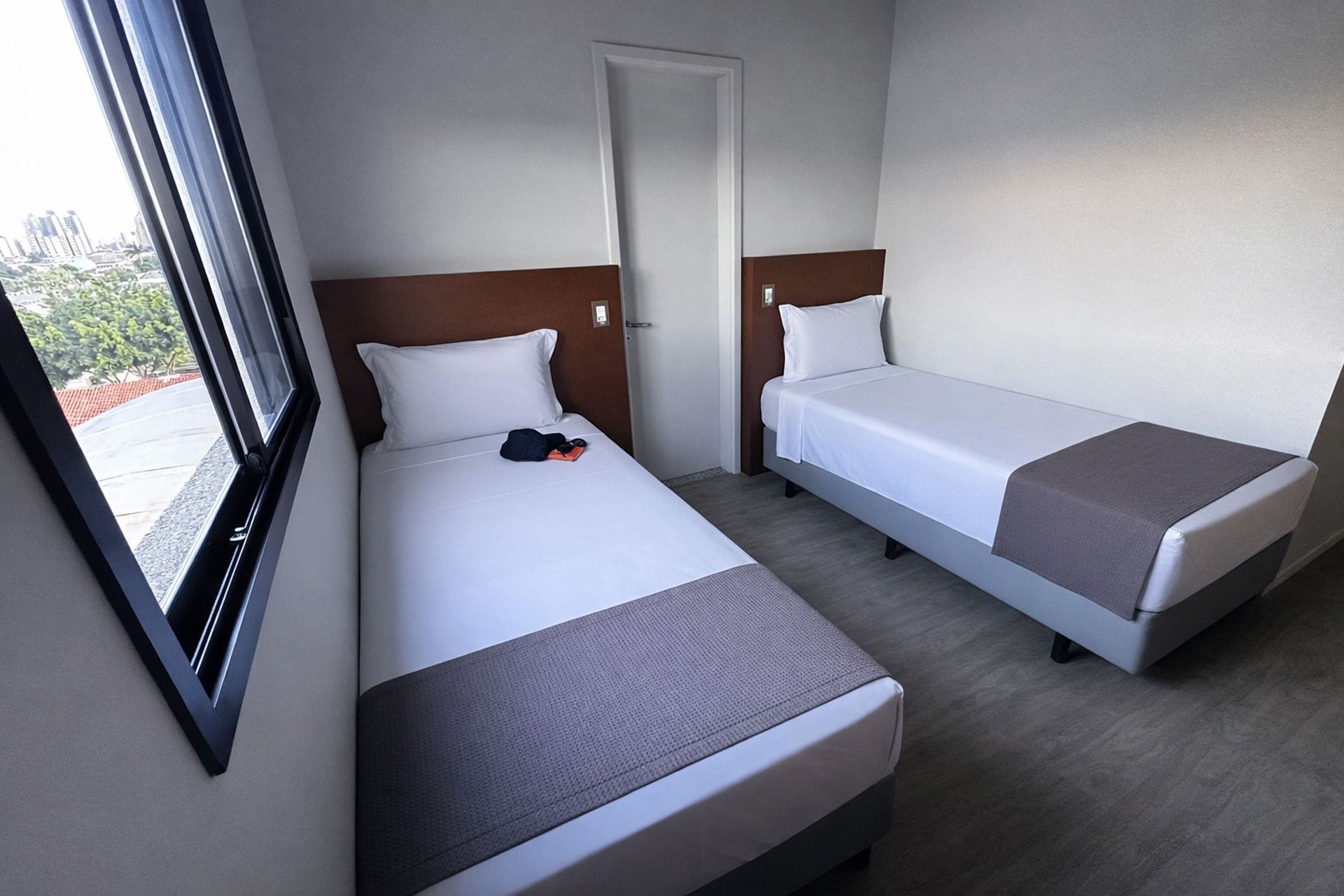 Duas camas de solteiro em um quarto de hotel bem iluminado. Lençóis brancos, peseiras cinza e uma janela.
