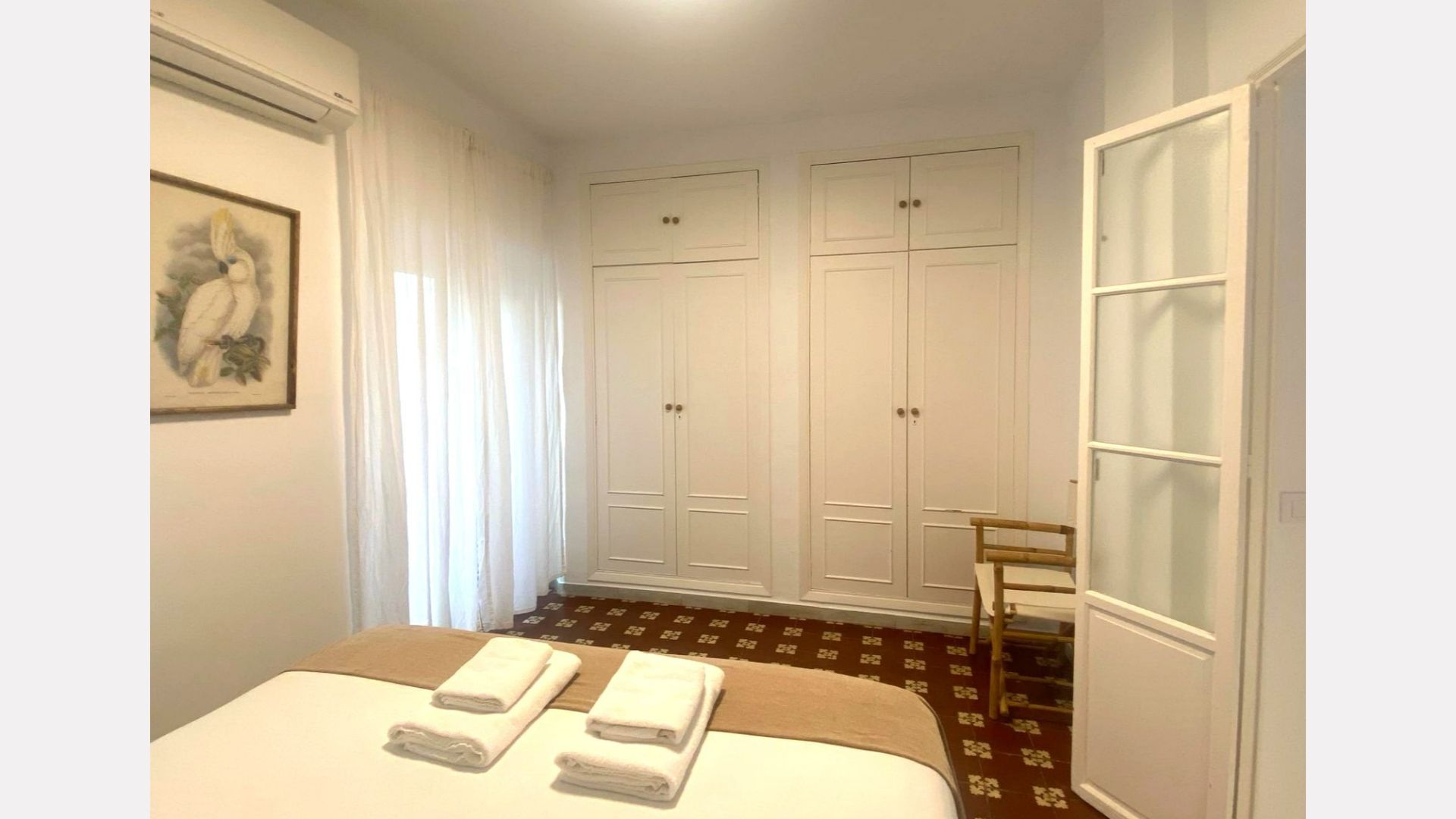 Bedroom with white cabinets, sheer curtains, bed with towels, artwork, and patterned floor.