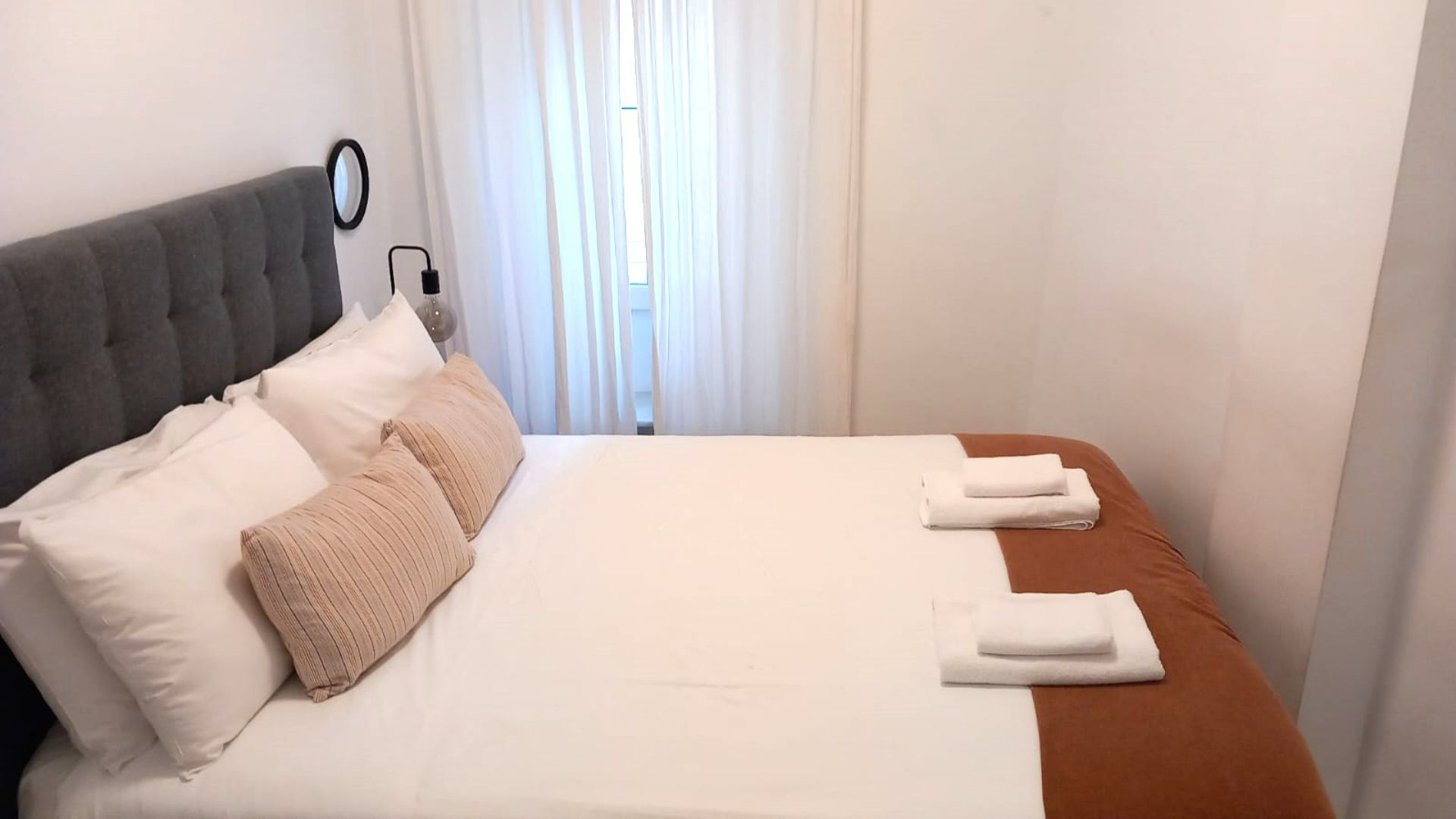 Bedroom with a bed made with white and brown linens, and pink pillows. White curtains in the background.