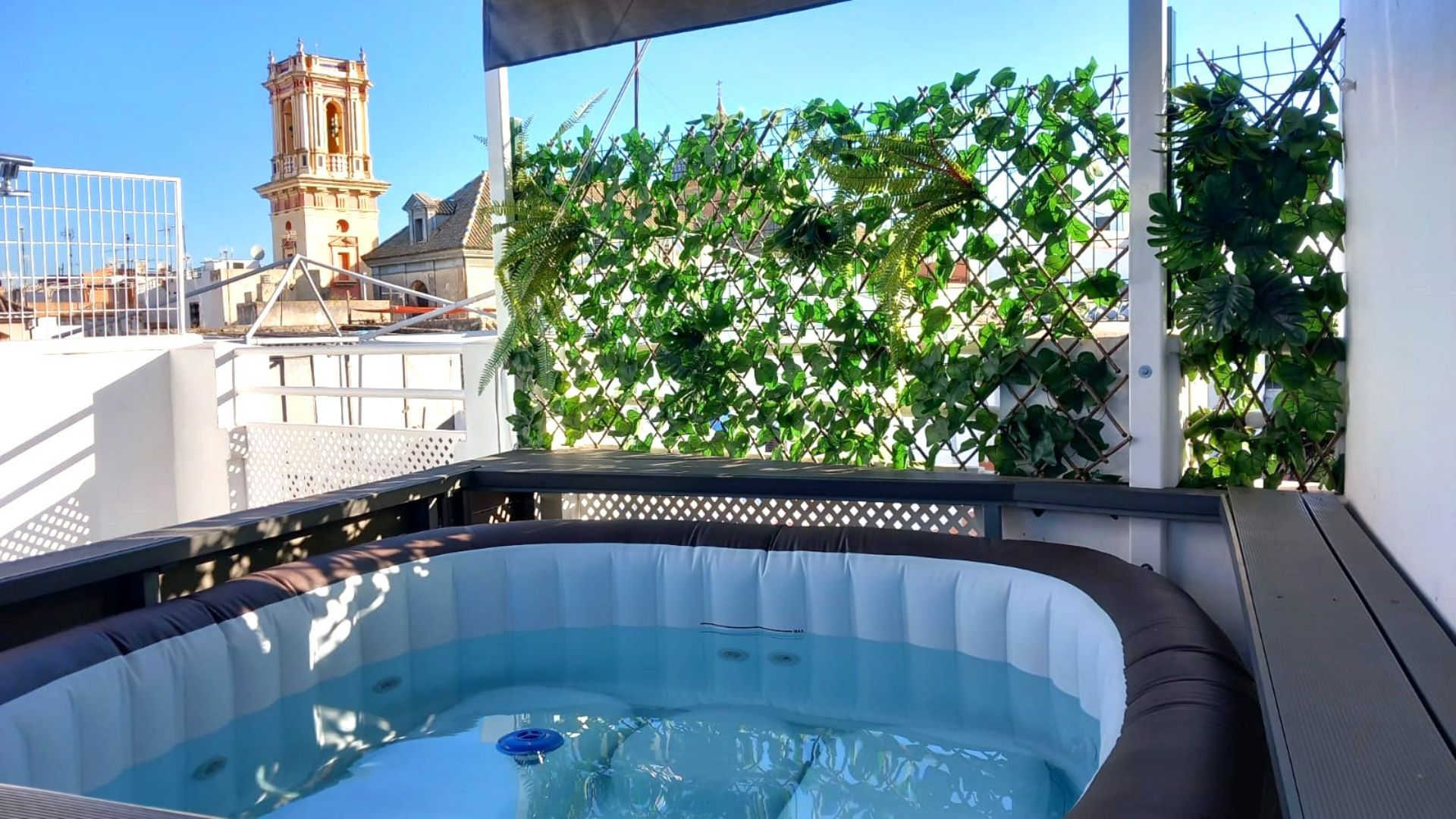 Rooftop hot tub with city view, green privacy screen, and clear blue sky.