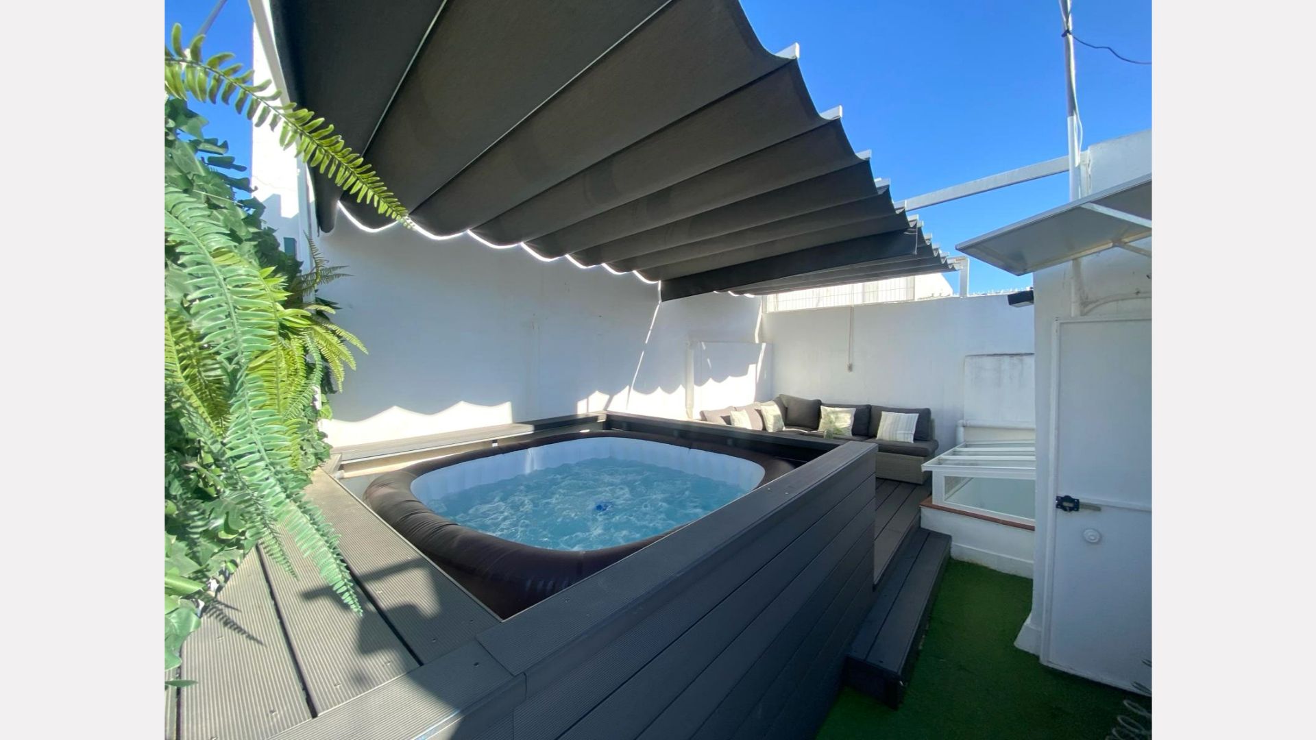Rooftop deck with hot tub, seating, and retractable awning under blue sky.
