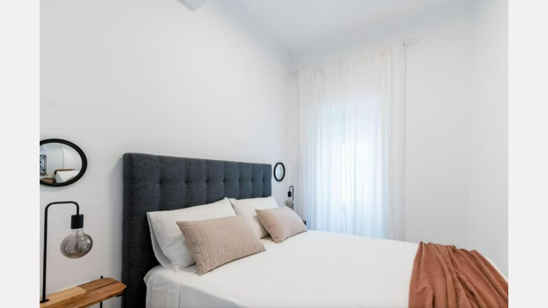 Bedroom with a gray upholstered headboard, white bedding, and peach blanket. A window with sheer curtains is visible.