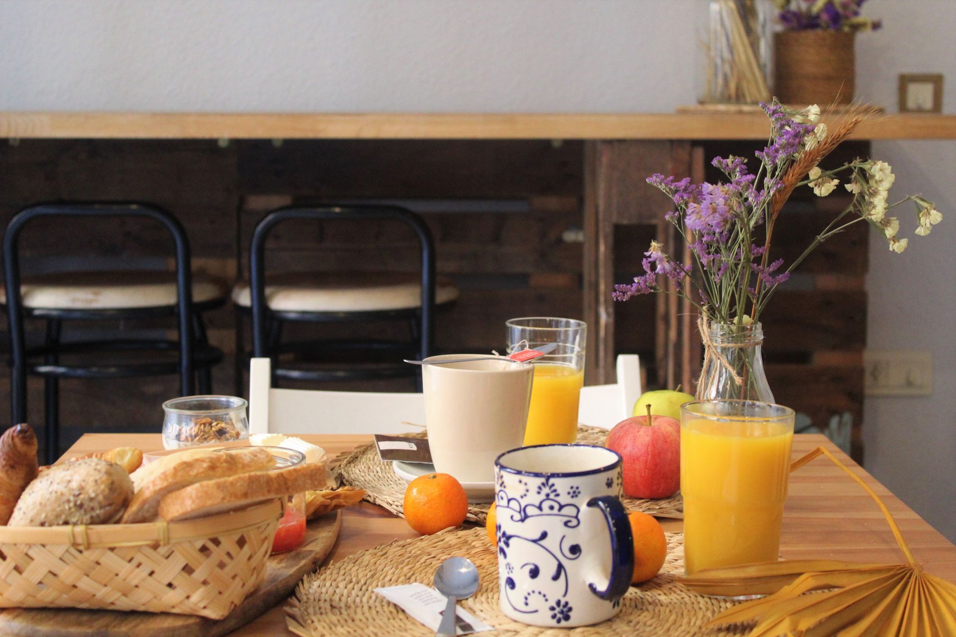 Breakfast table set with bread, fruit, and orange juice; flowers in a vase; chairs in the background.