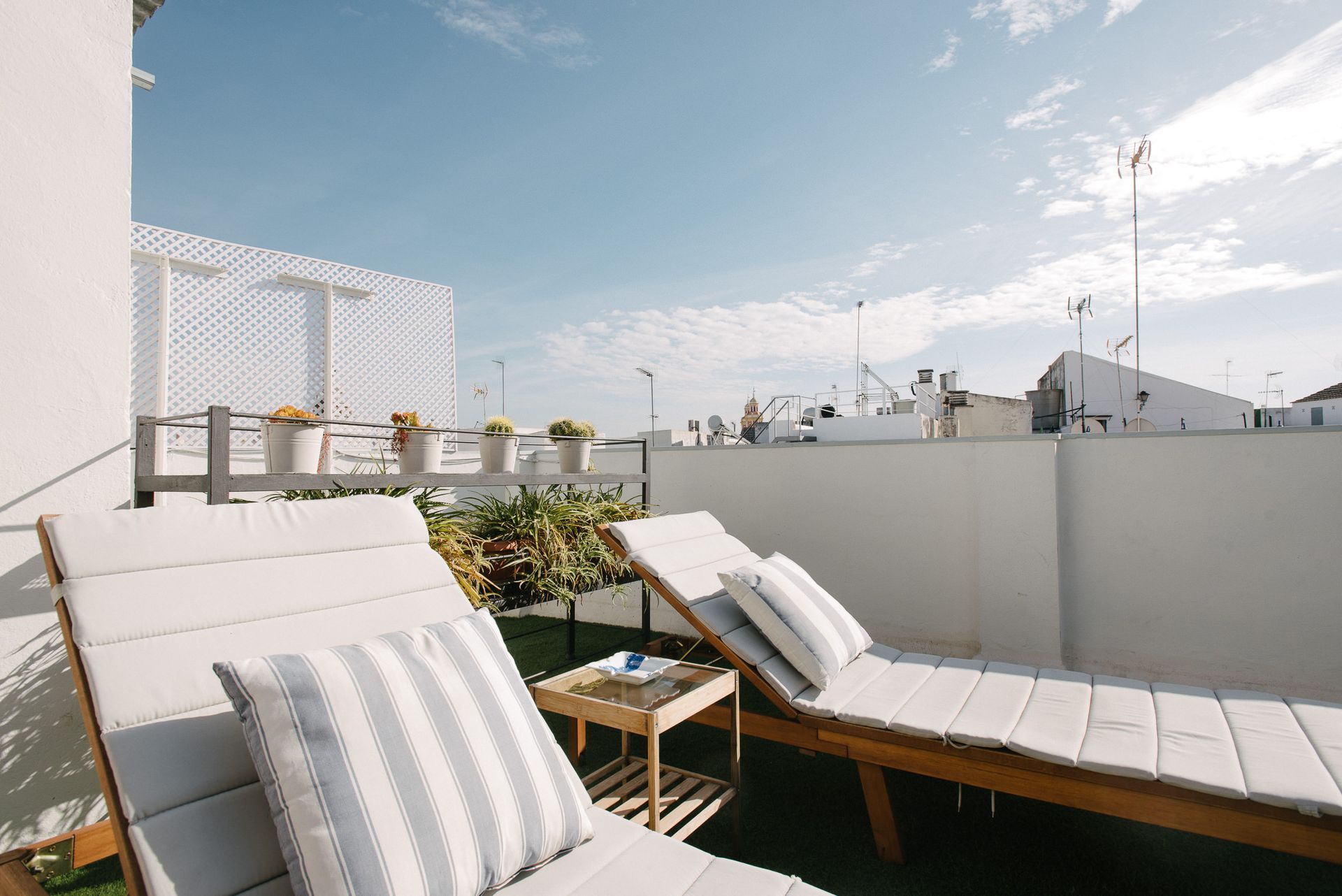 Two lounge chairs on a rooftop terrace with blue sky, white walls, and potted plants.